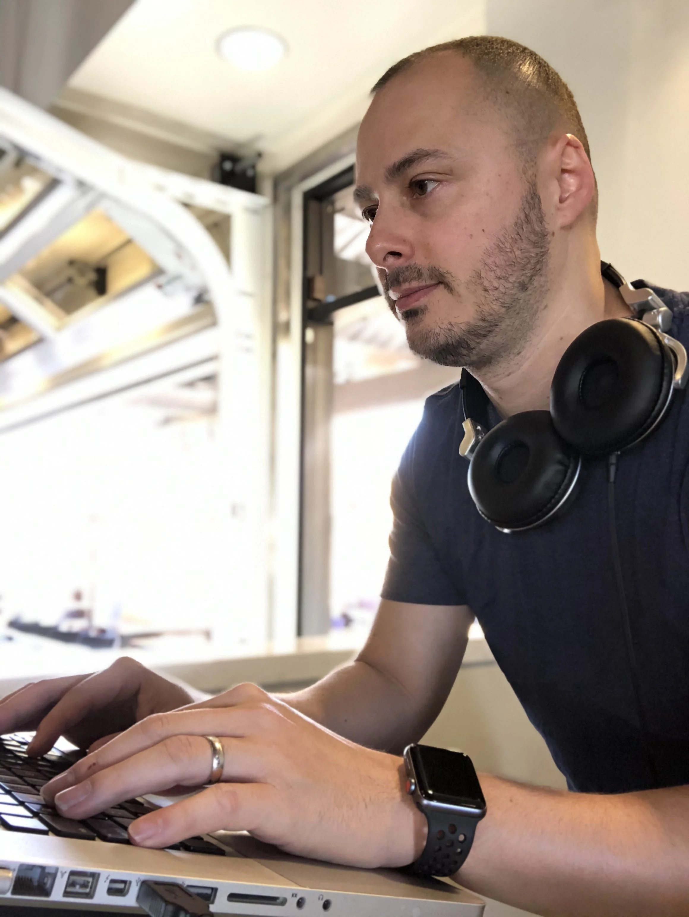 DJ MV wearing a wristwatch and headphones around his neck, is working on a laptop. He is sitting indoors near a window, with a focused expression.