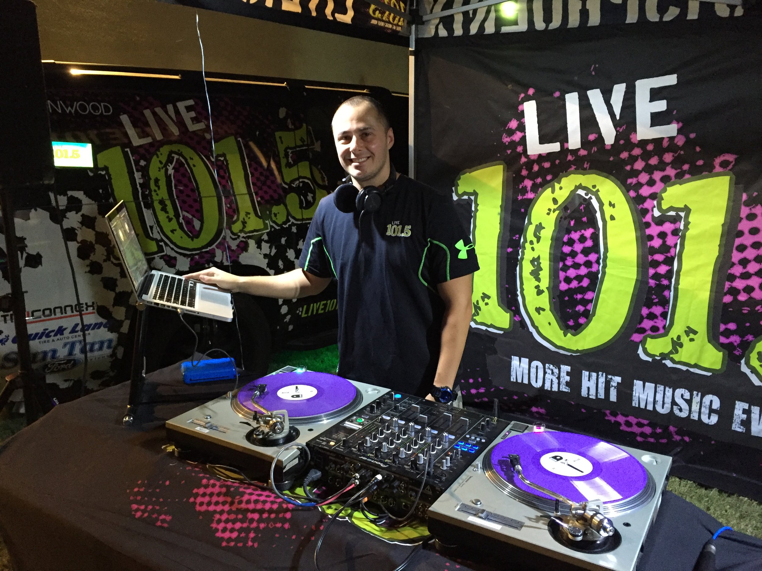 A man standing behind DJ equipment at a radio station event with a banner in the background that reads 'Live 101.5 more hit music everywhere.' He is smiling, wearing a dark shirt with the station's logo, and has headphones around his neck.
