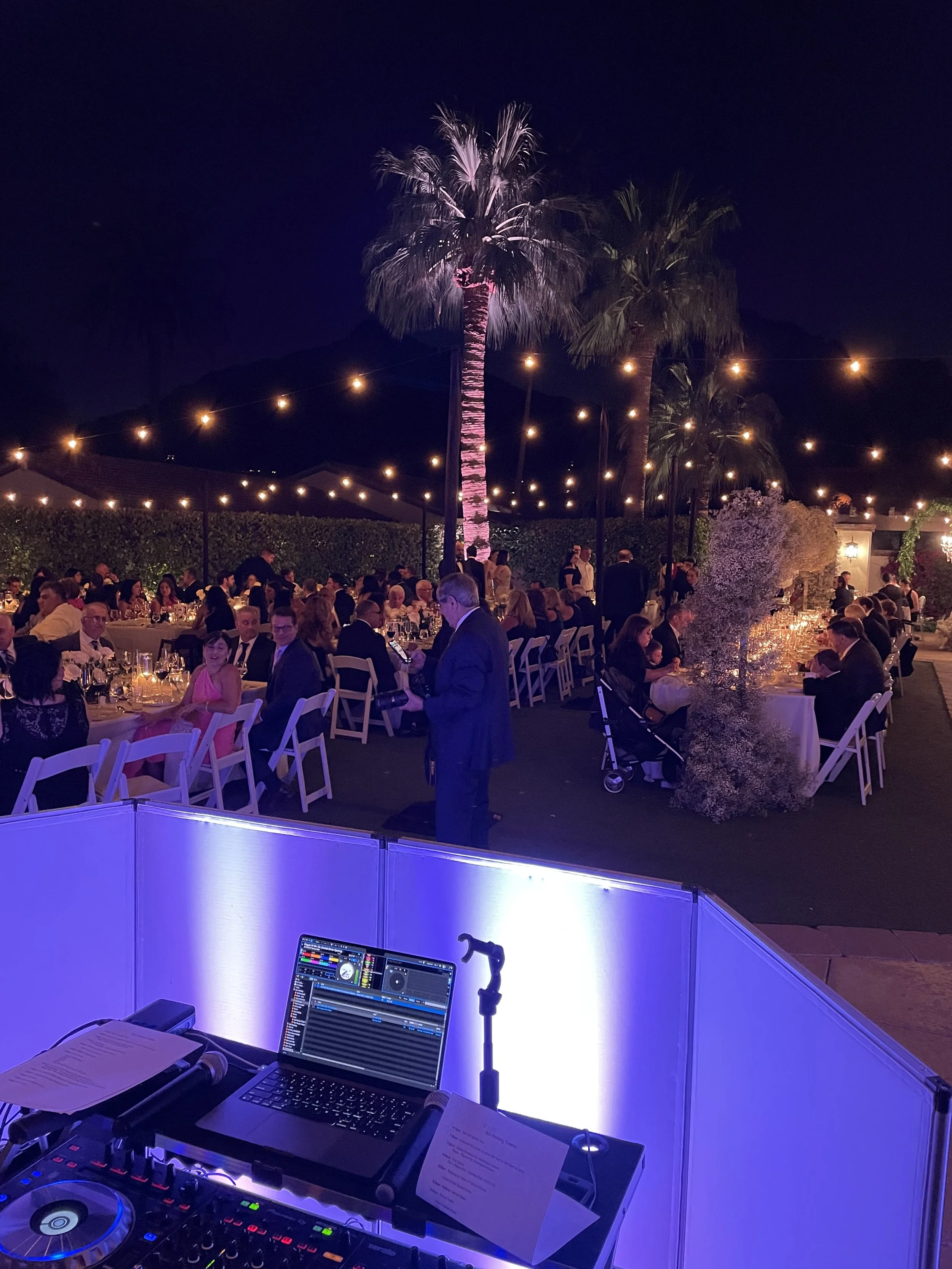Nighttime outdoor event with string lights, palm trees, and a large gathering of people seated at long tables, with a DJ setup in the foreground.