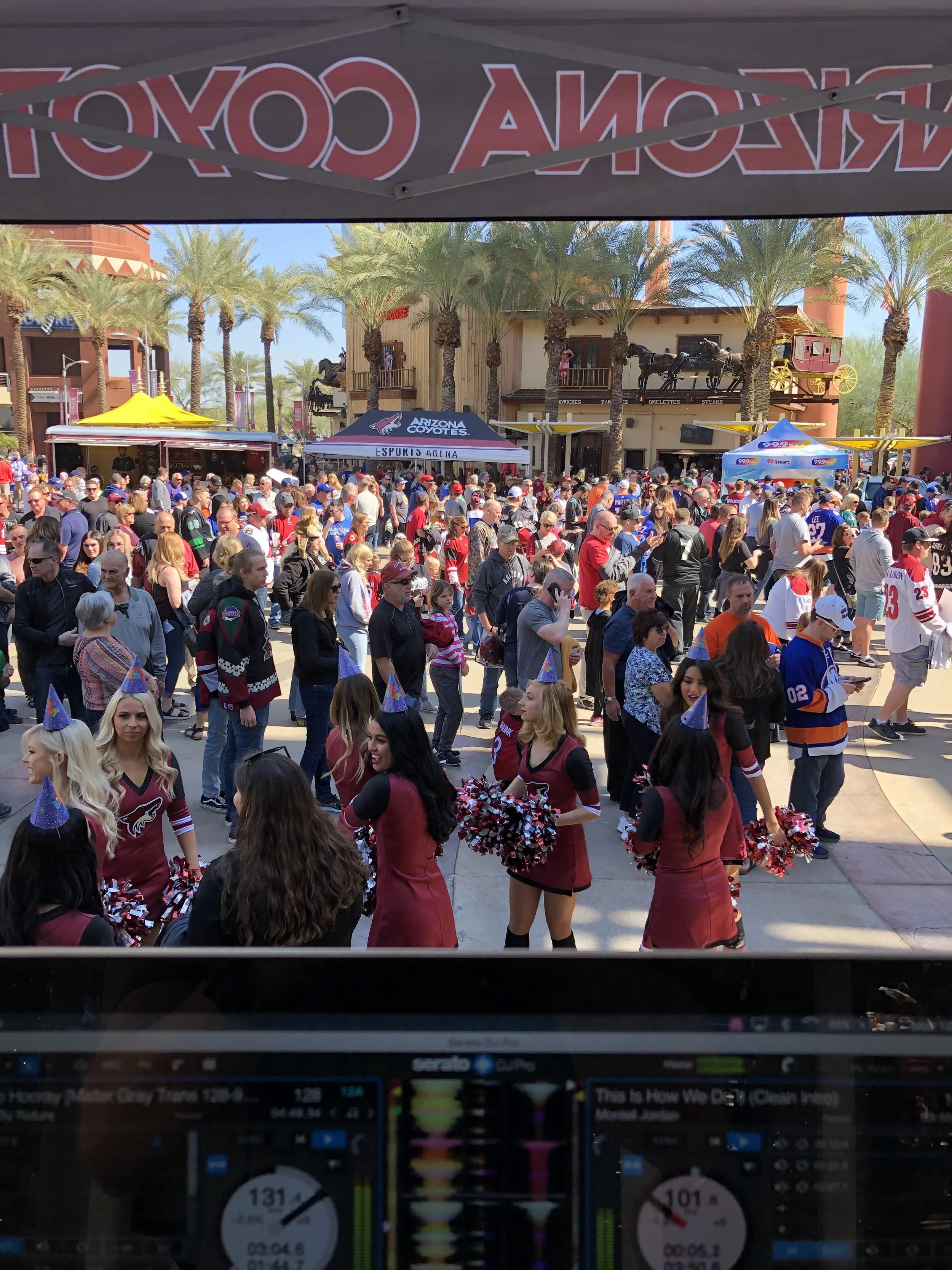 Crowd of people at a Arizona Coyotes event outdoors, with cheerleaders in front wearing red and black uniforms, and vendor tents in the background under palm trees and a clear sky.