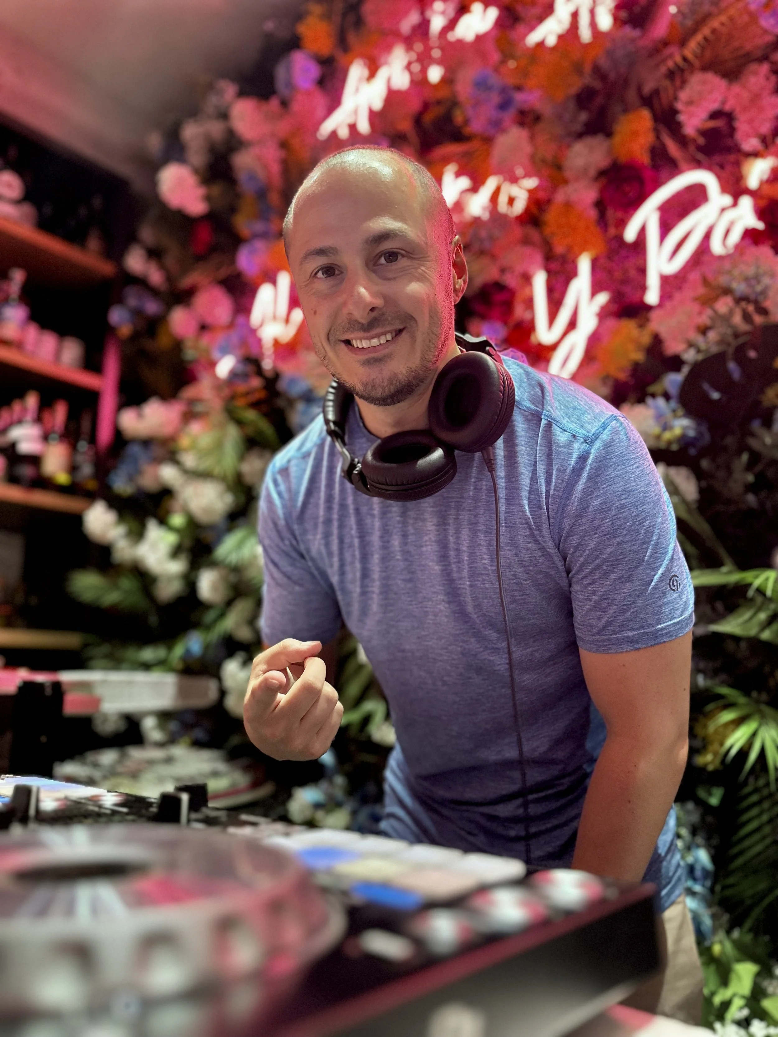 DJ MV with a headphone around his neck, smiling and making a fist gesture, standing in front of a colorful floral background with neon signs.