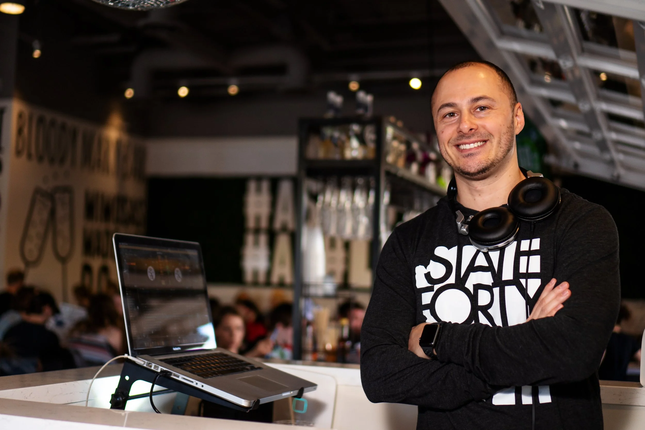 standing with arms crossed in a modern, well-lit cafe or coworking space. He wears a black long-sleeved shirt with white graphic text, headphones around his neck, and a smartwatch on his wrist. A laptop on a stand is in front of him, with a group of people in the background.