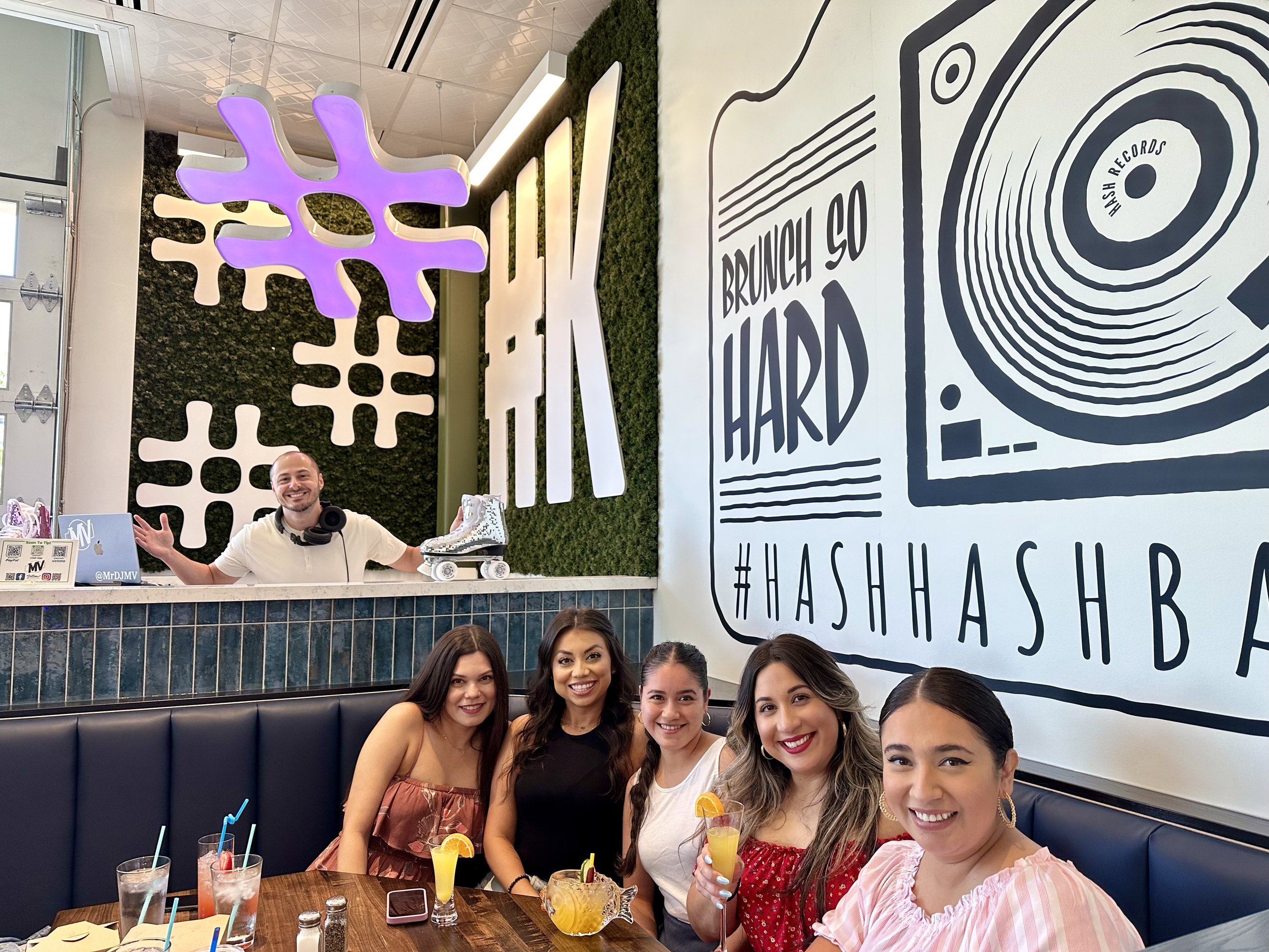 Five women sitting at a table with drinks, with a DJ MV in the background and large hashtag symbols and graphics on the wall.