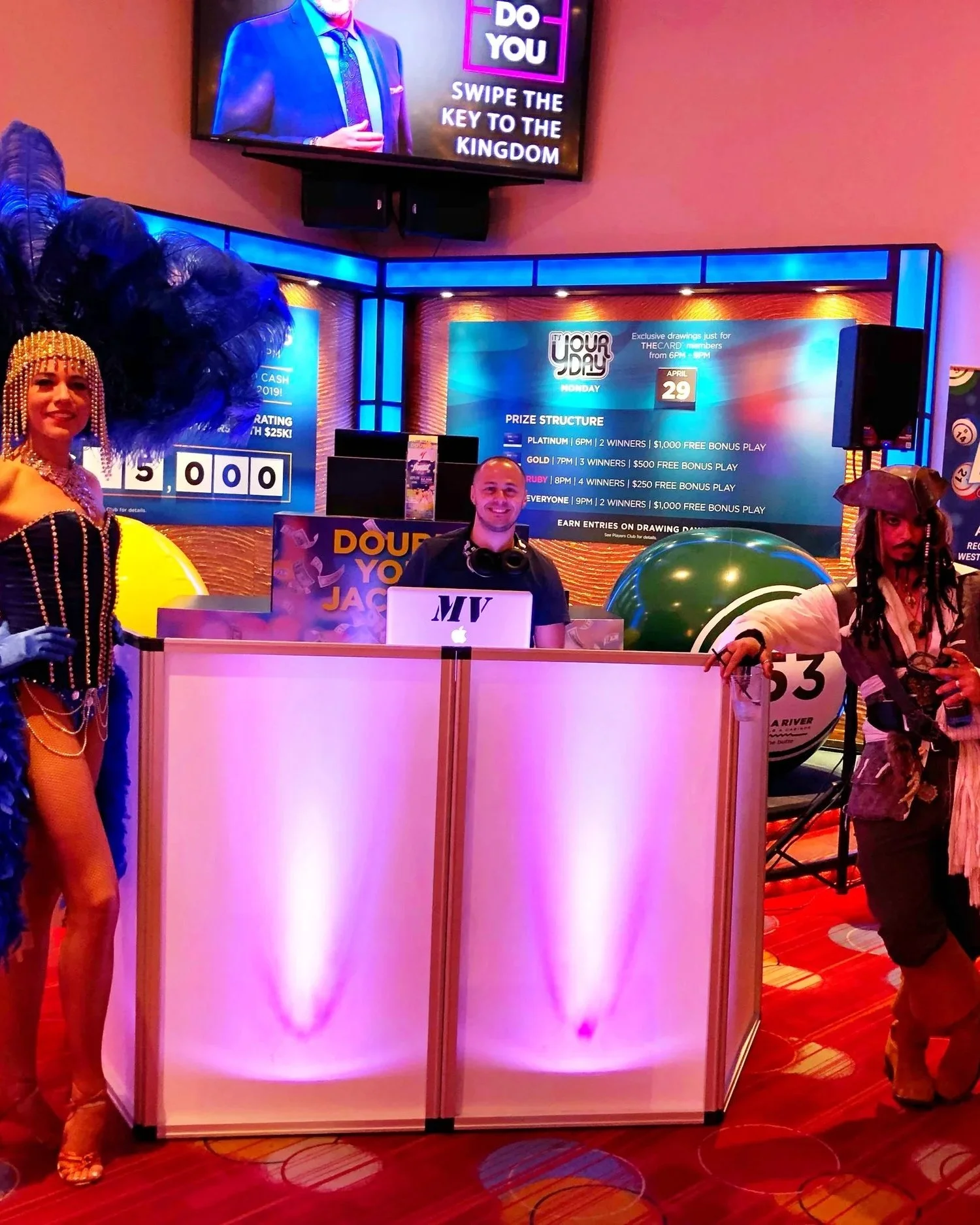 DJ MV sitting in a DJ booth with a smile, surrounded by two women in costume costumes at a lively event, with digital screens displaying promotional information and colorful lighting in the background.