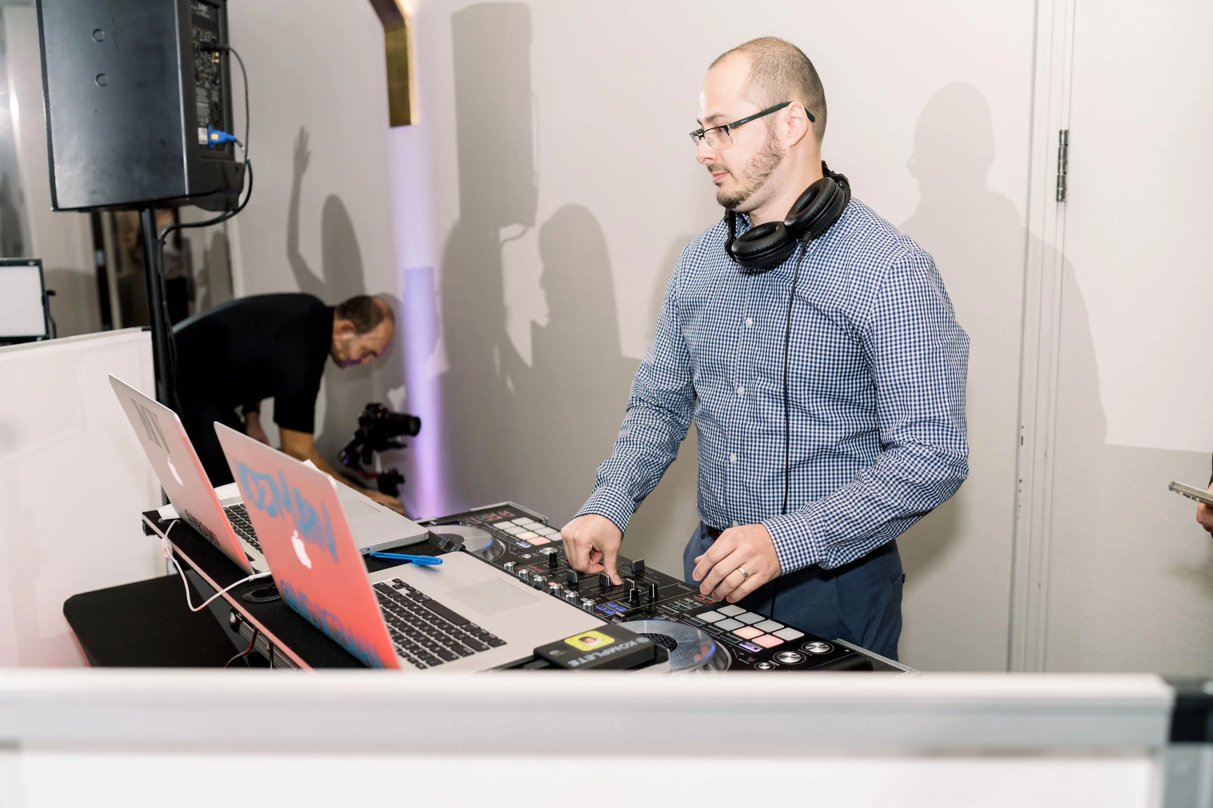 DJ MV mixing music at a party with laptops and equipment, wearing glasses and headphones around his neck.