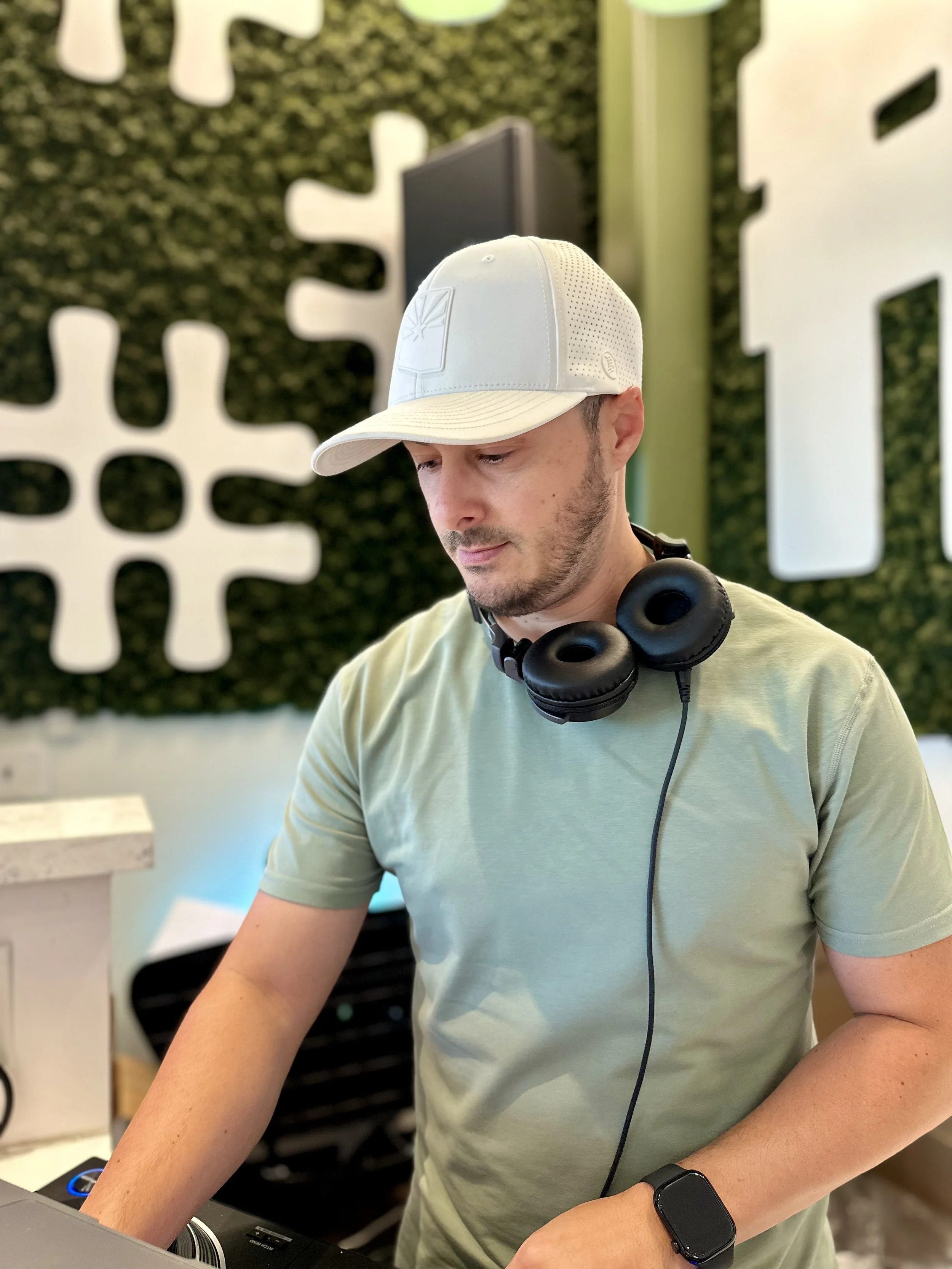DJ MV wearing a white cap, black headphones around his neck, and a light green t-shirt, working at a DJ booth.