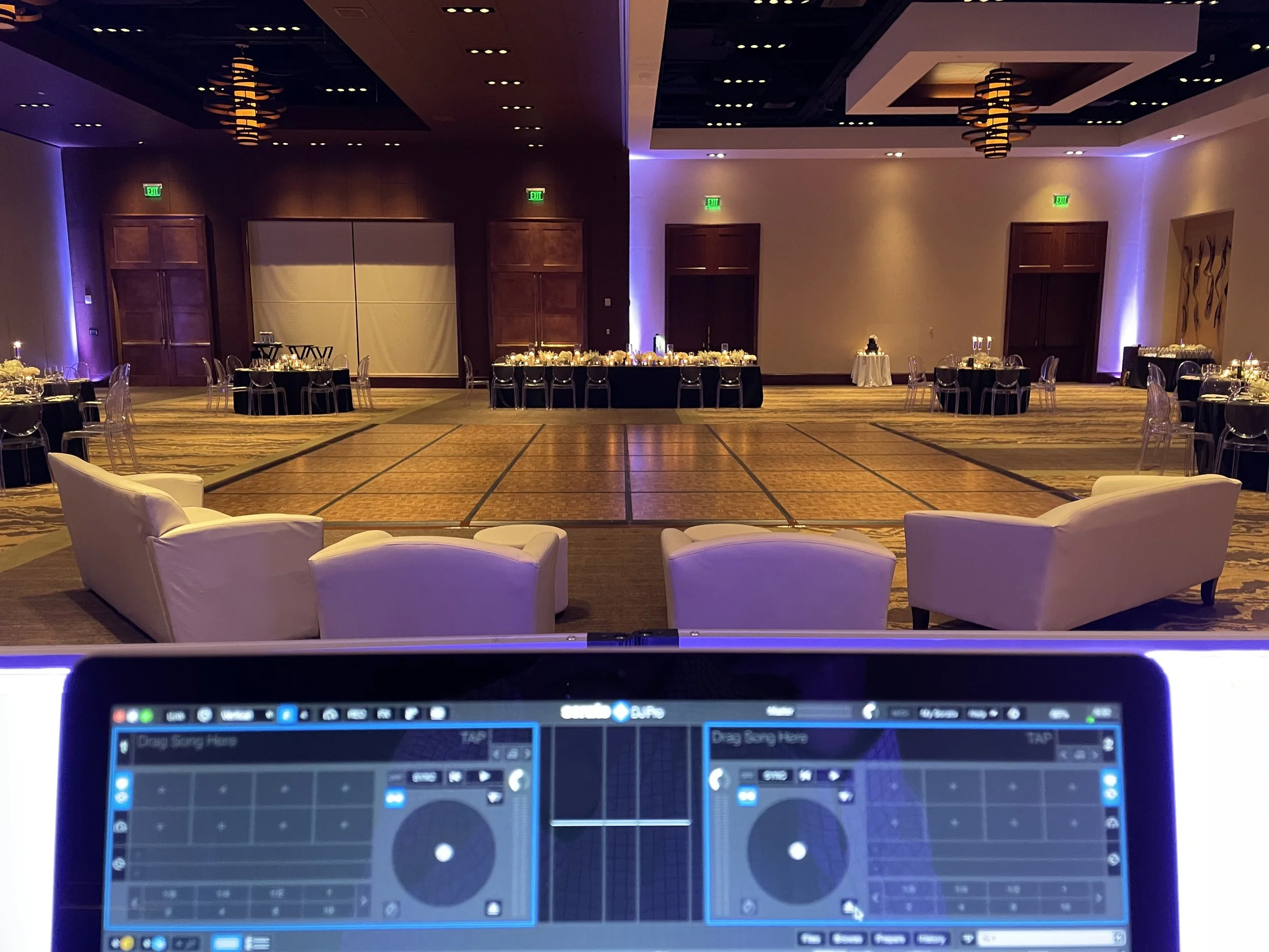 Empty banquet hall with round tables set for an event, decorated with candles and flowers, and empty chairs around them. Two white armchairs are in the foreground facing the hall, and a DJ mixing console is visible at the bottom of the image.