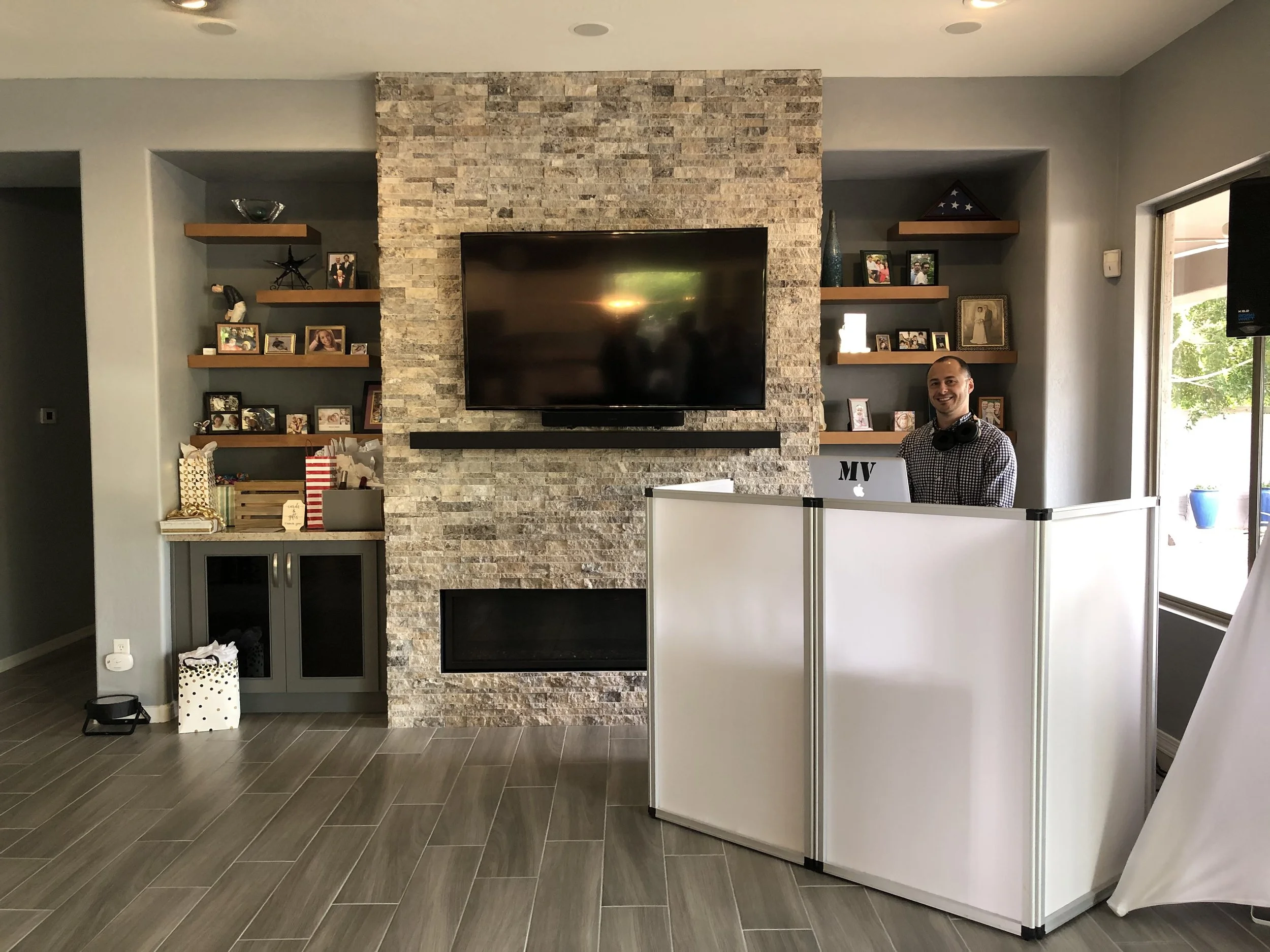 DJ MV standing behind a white podcast booth with headphones around his neck, in a living room with a stone fireplace, a mounted TV, and corner shelves filled with photos and decor.