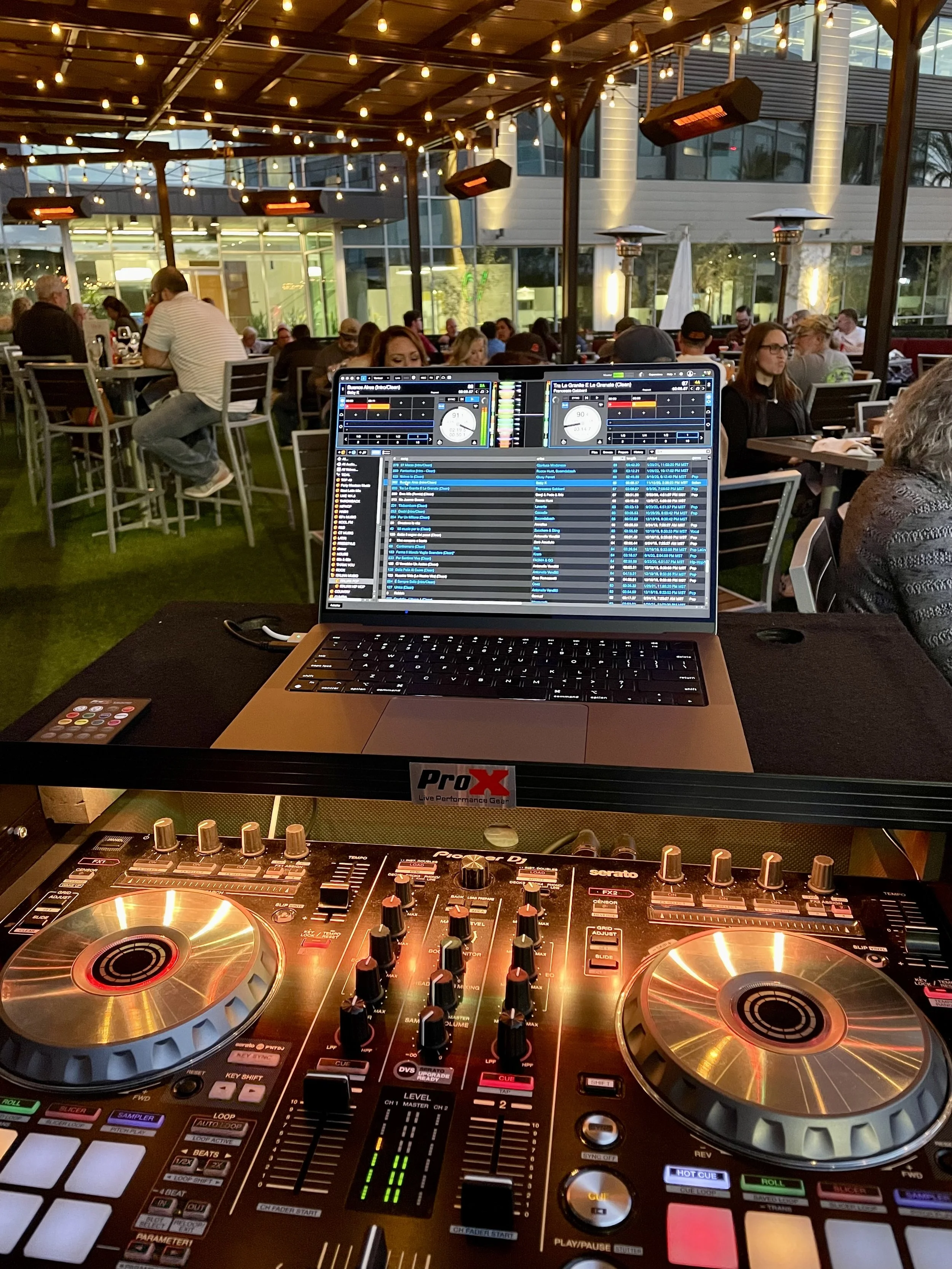 A DJ setup with a laptop and turntables in a lively outdoor venue with people dining and socializing under string lights.