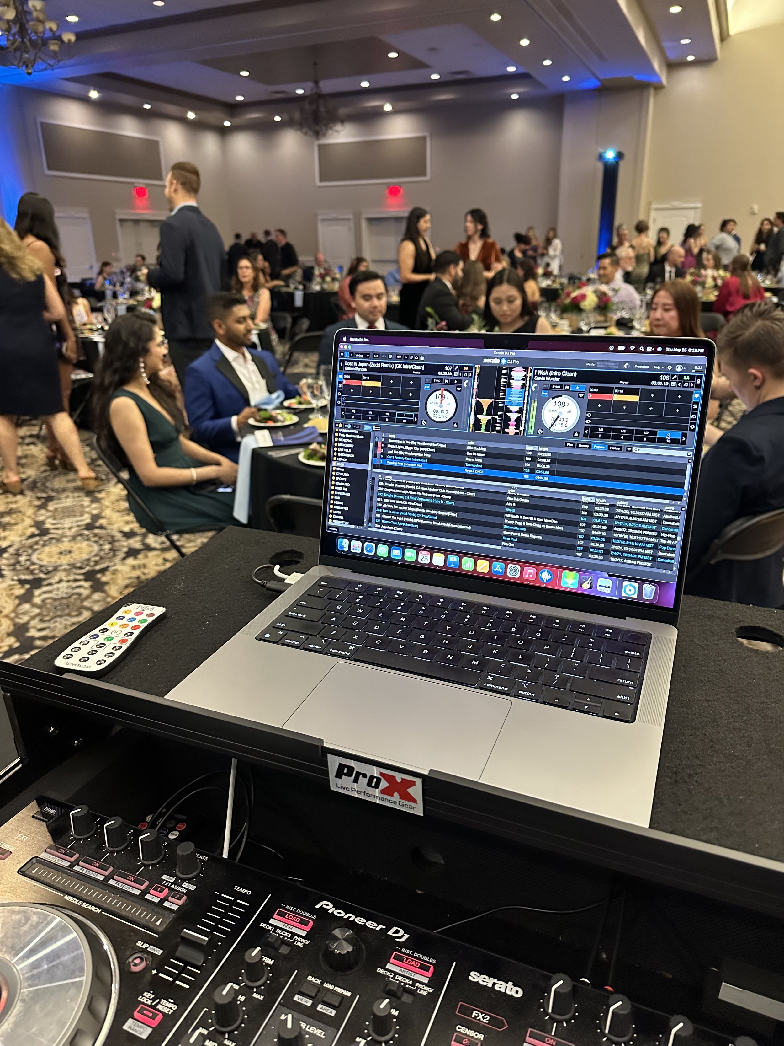 A DJ setup with a laptop and mixing equipment, overlooking a banquet hall filled with guests seated at tables during a formal event or celebration.