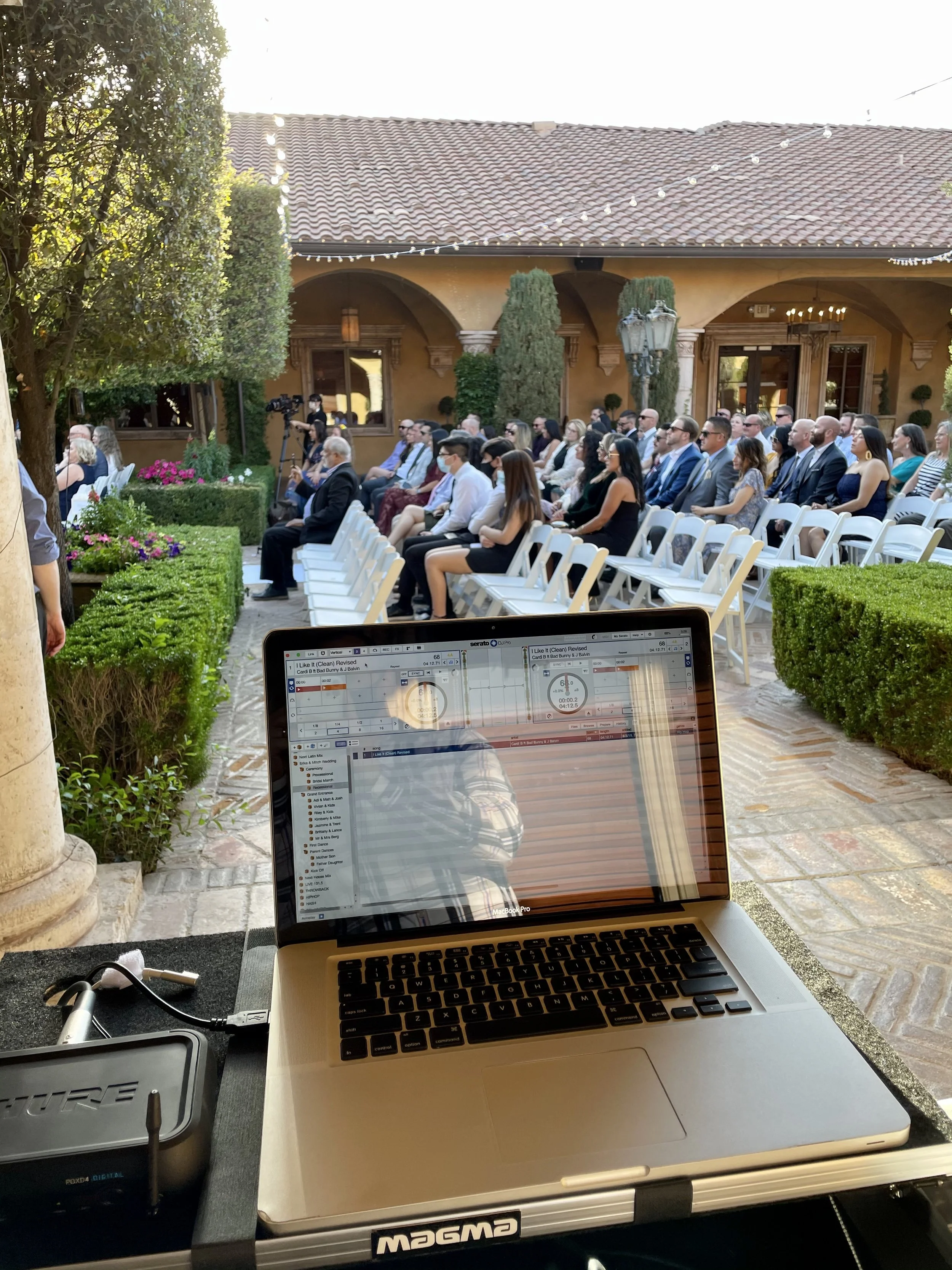 Outdoor wedding ceremony with seated guests facing the altar, taken from behind a DJ setup with a laptop showing a music playlist.