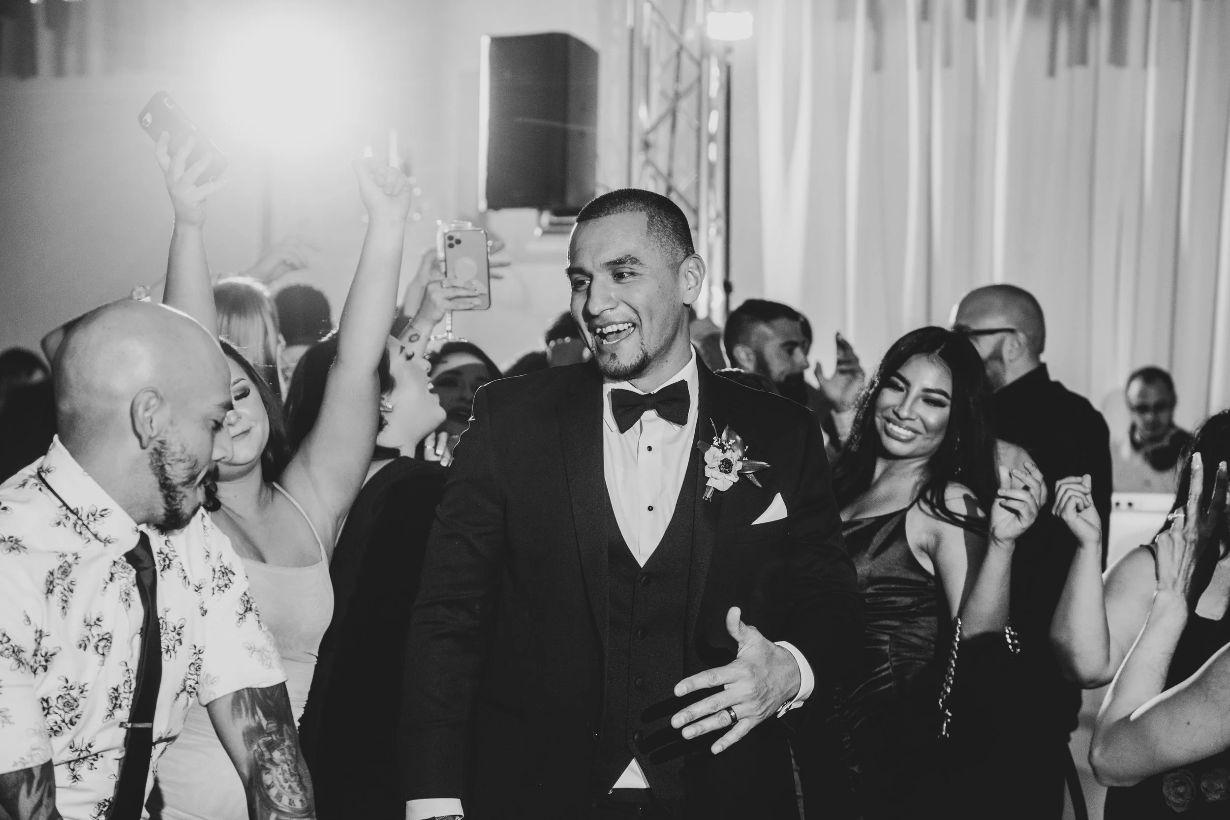 A man in a tuxedo with a bow tie and boutonniere is smiling at a celebration or party. He is surrounded by other smiling and dancing people, some taking photos with their phones.