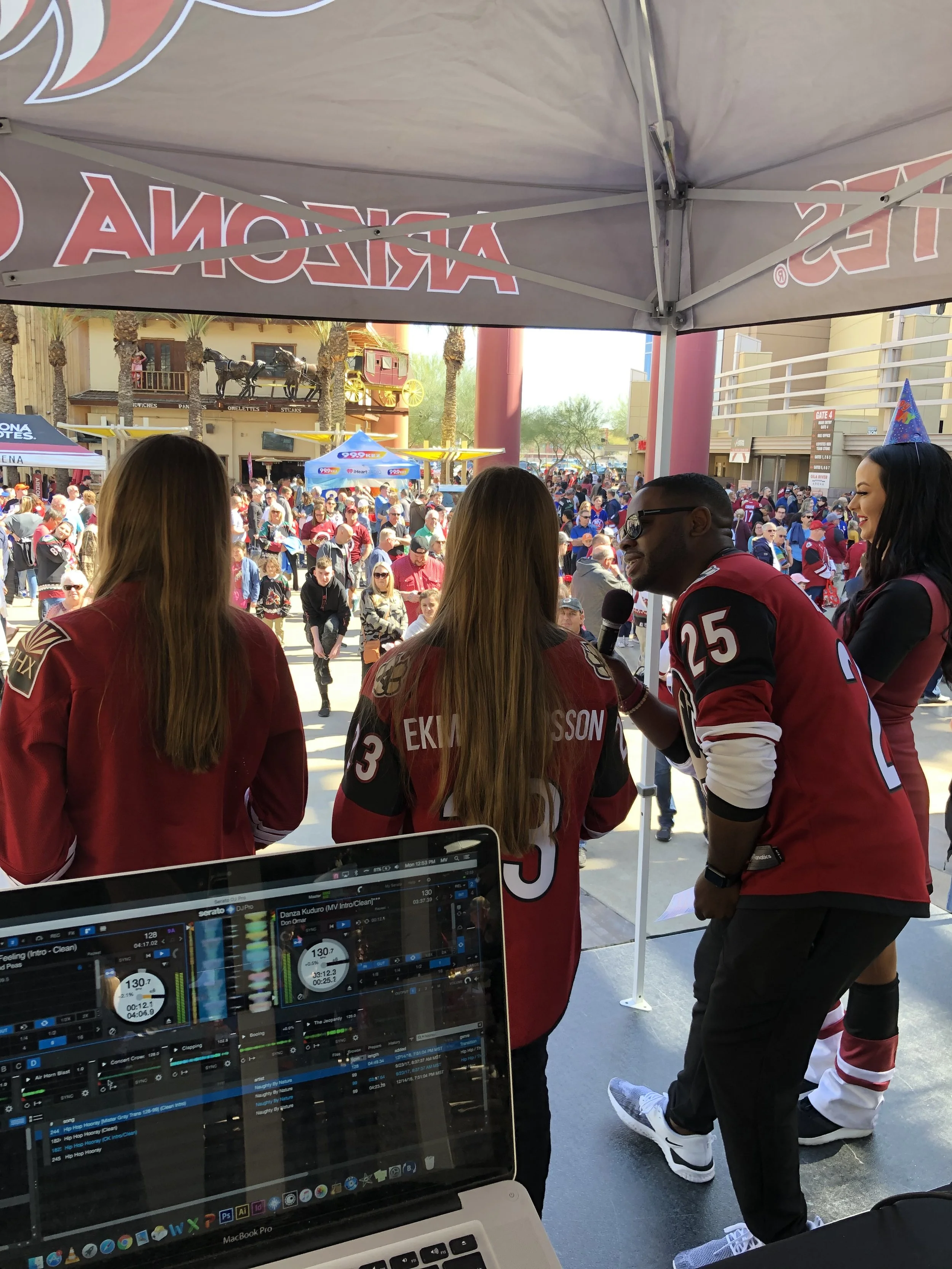 A crowd of people gathered outdoors, with some wearing hockey jerseys, and a man holding a microphone engaging with the crowd. There are tents and buildings in the background.