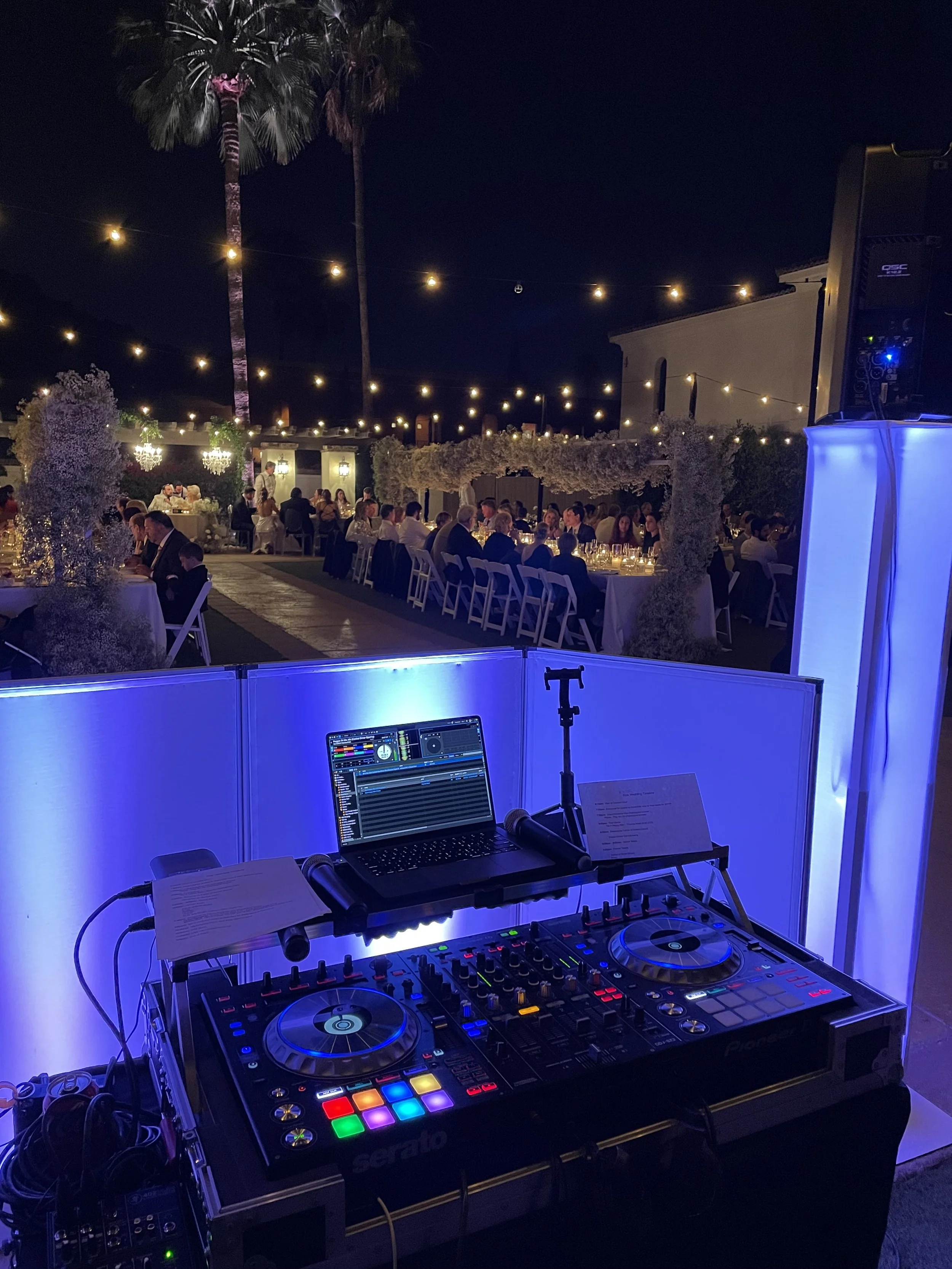 DJ equipment set up outdoors at night with a laptop and colorful lights, overlooking a decorated outdoor dining area filled with people under string lights and palm trees.