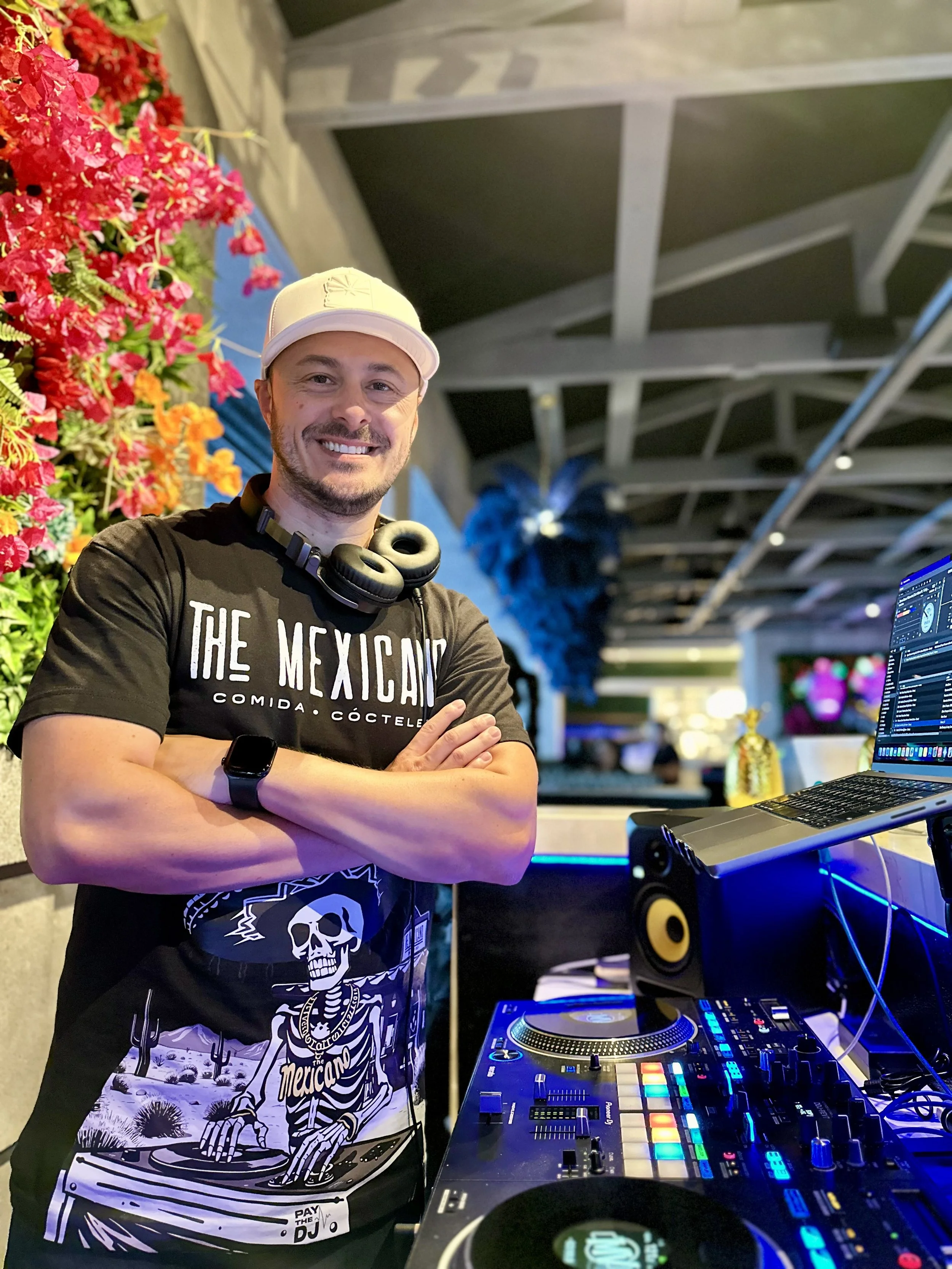 DJ MV wearing a white cap, black t-shirt with the words 'The Mexicano' and a skeleton graphic, and a black watch stands with arms crossed behind DJ equipment in a decorated venue with colorful flowers and blue feather decorations.