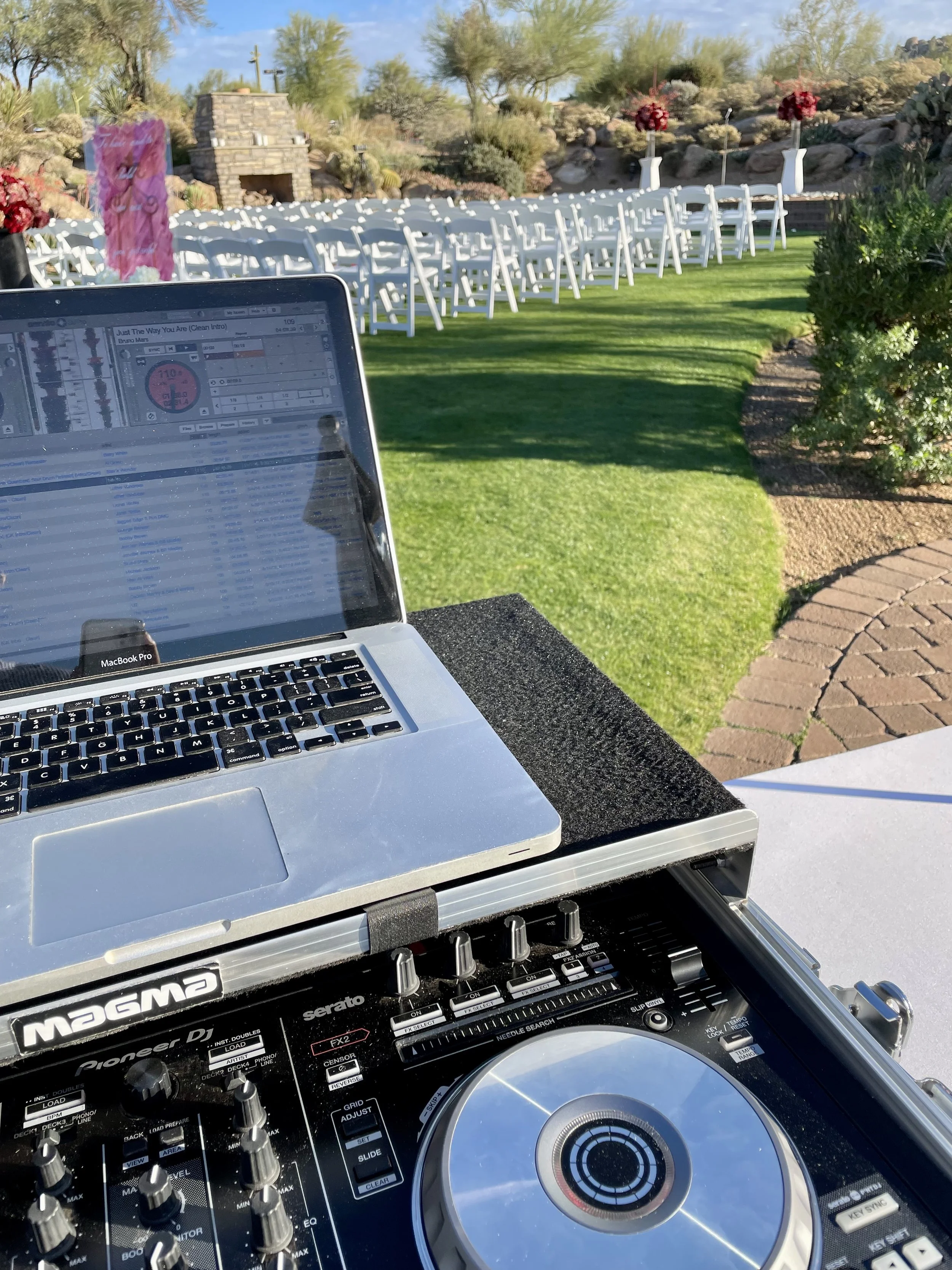 DJ equipment set up outdoors on a table with a MacBook and a Pioneer DJ controller, overlooking rows of white chairs and floral arrangements at a sunny outdoor event space with desert landscaping.