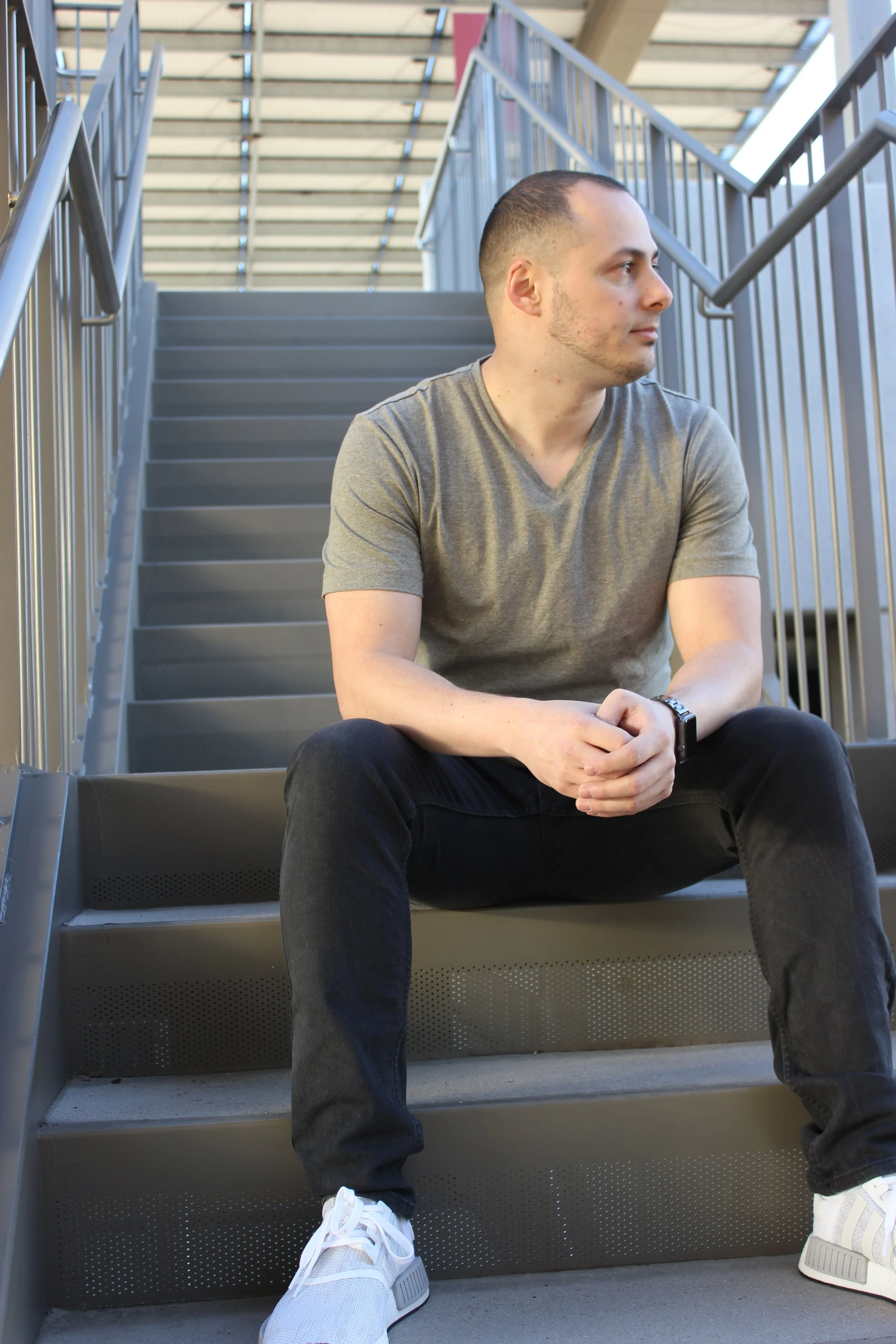 DJ MV wearing a grey t-shirt, black jeans, and white sneakers sitting on outdoor metal stairs, looking to the right.