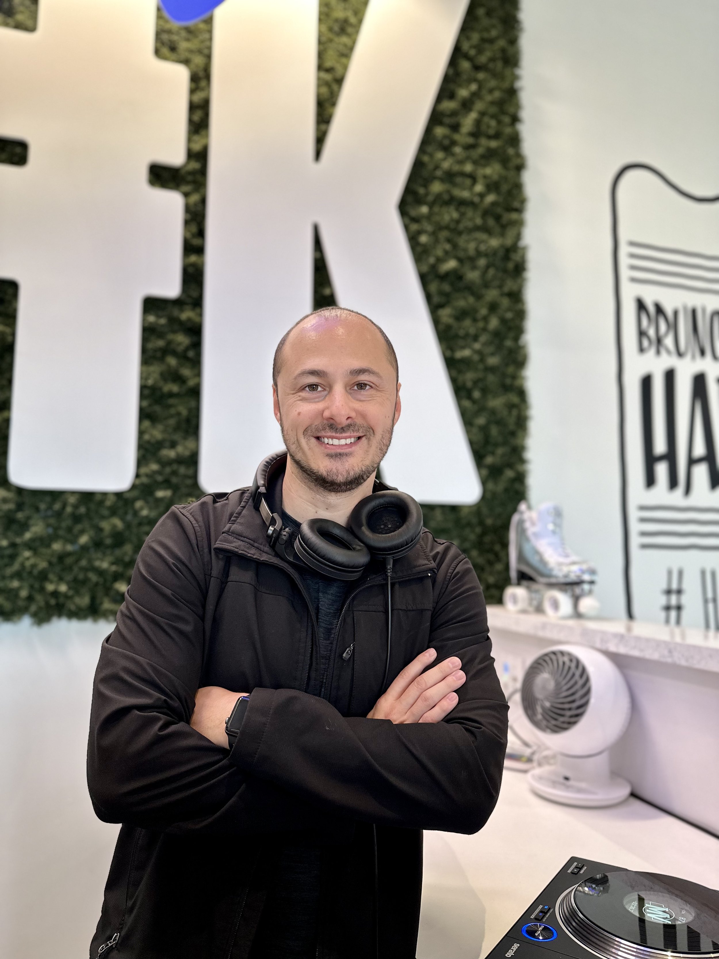 DJ MV posing, wearing a black jacket and headphones around his neck, is smiling at the camera with arms crossed, standing in front of a wall with large white lettering and a graphical mural at Hash Kitchen Gilbert.