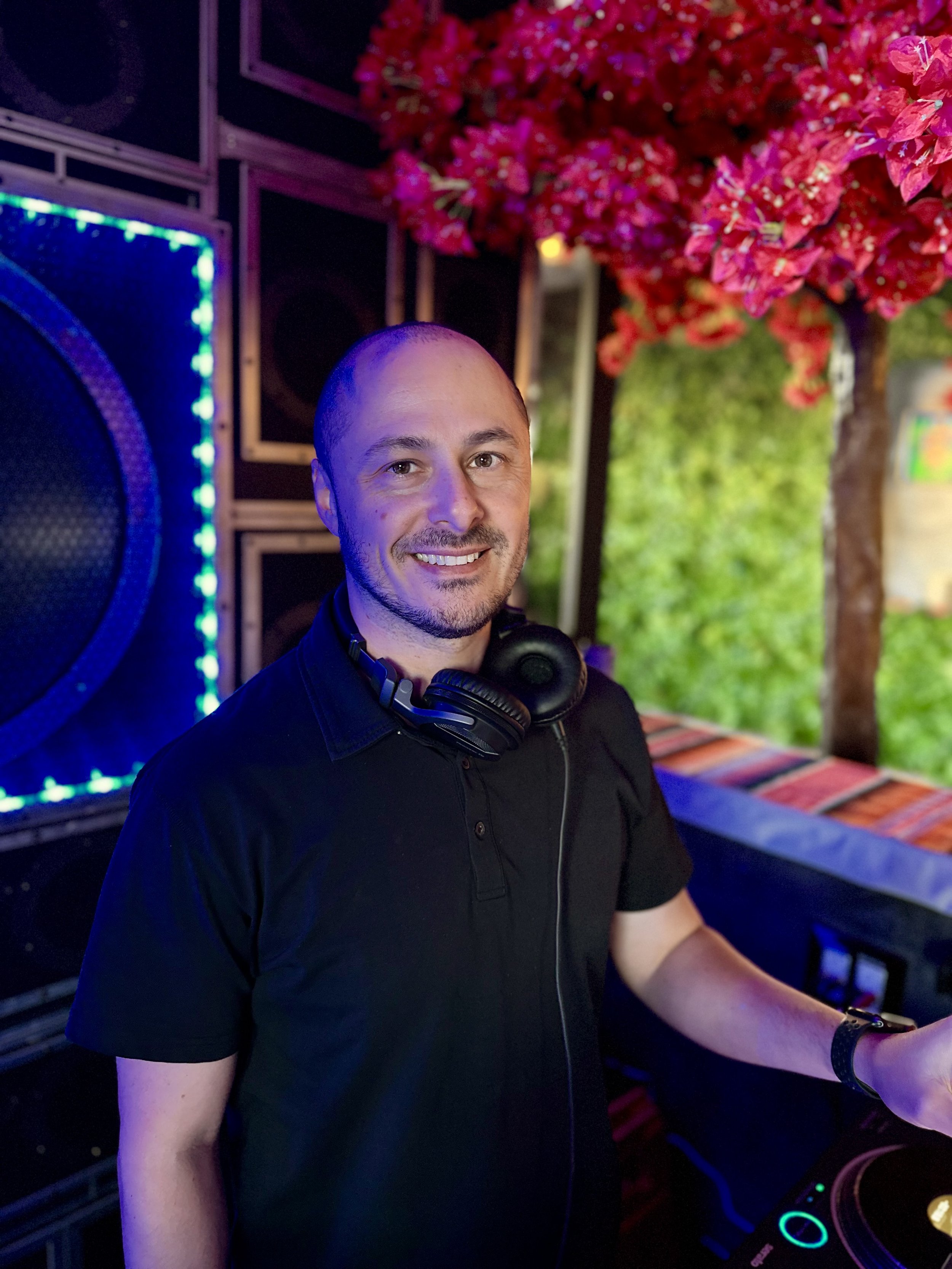 DJ MV wearing a black polo shirt and headphones around his neck, standing behind a DJ mixer in a decorated venue with colorful lighting, a large speaker, a pink flowering tree, and green foliage in the background.