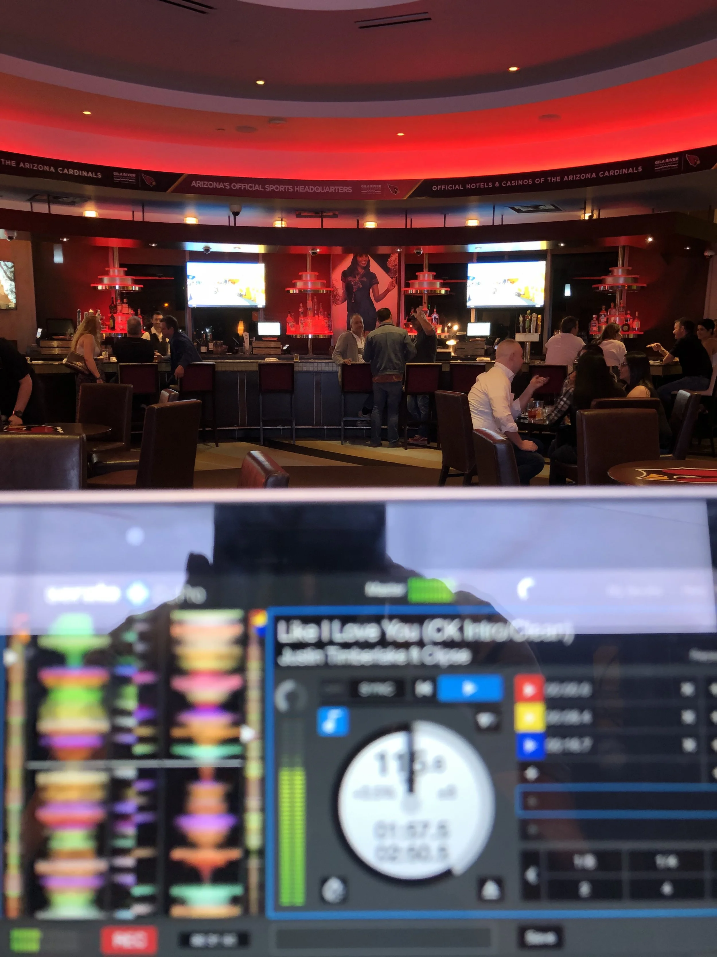 A bar scene with people socializing. The bar has a red and black color scheme, with illuminated shelves displaying bottles. Multiple televisions are mounted, and a large poster of a woman is behind the bar. The foreground shows DJ equipment with colorful lights and a digital timer.