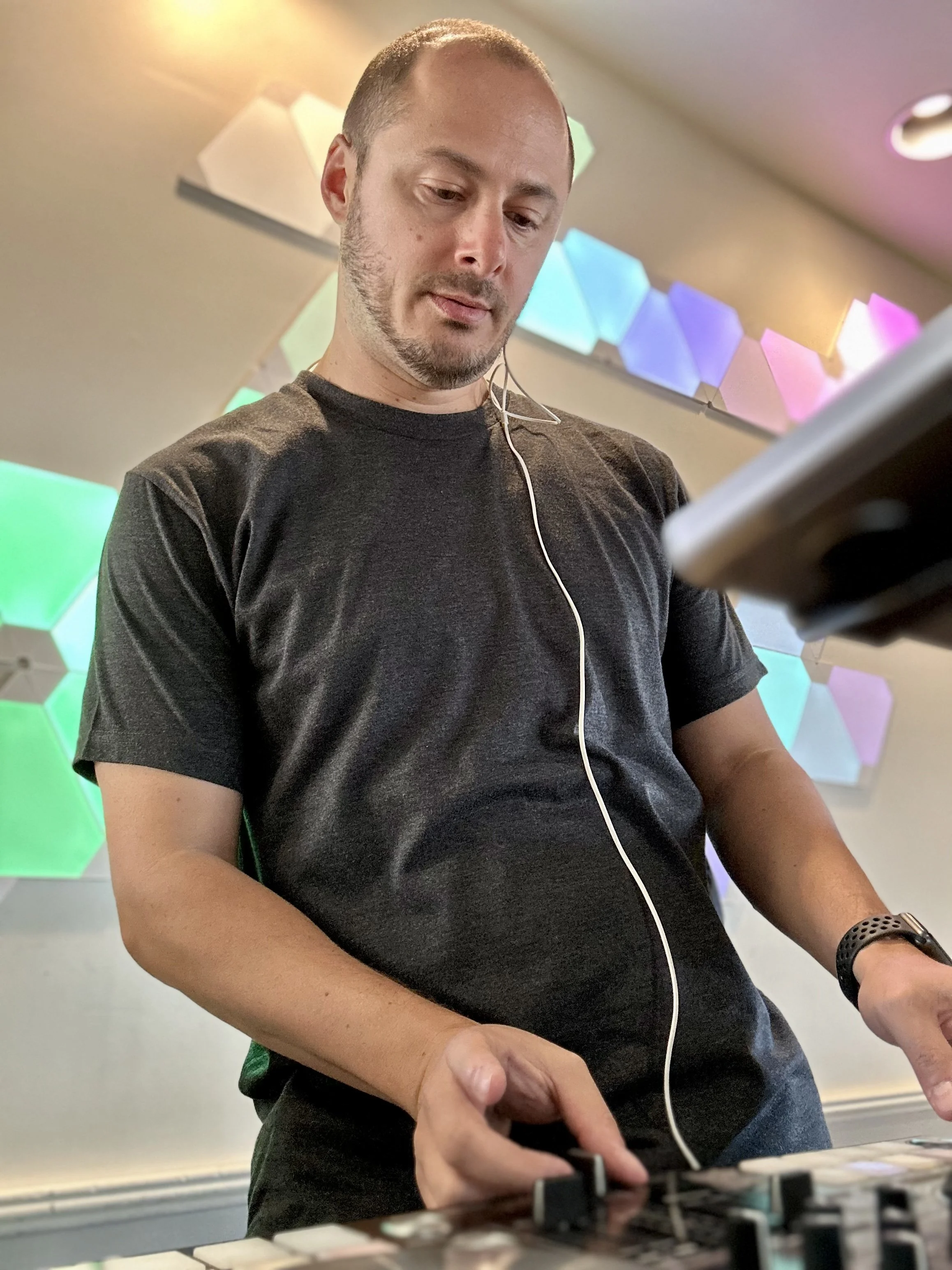DJ MV wearing a black t-shirt is DJing with a mixer. He has a white earphone in his ear and a black watch on his wrist. Behind him are colorful geometric wall panels illuminated in various colors.