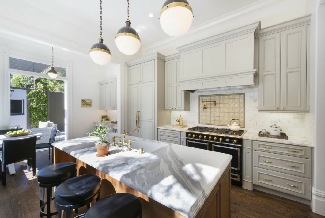 A modern kitchen with white cabinets, a marble island, black and gold appliances, pendant lighting, and a dining area with outdoor access.