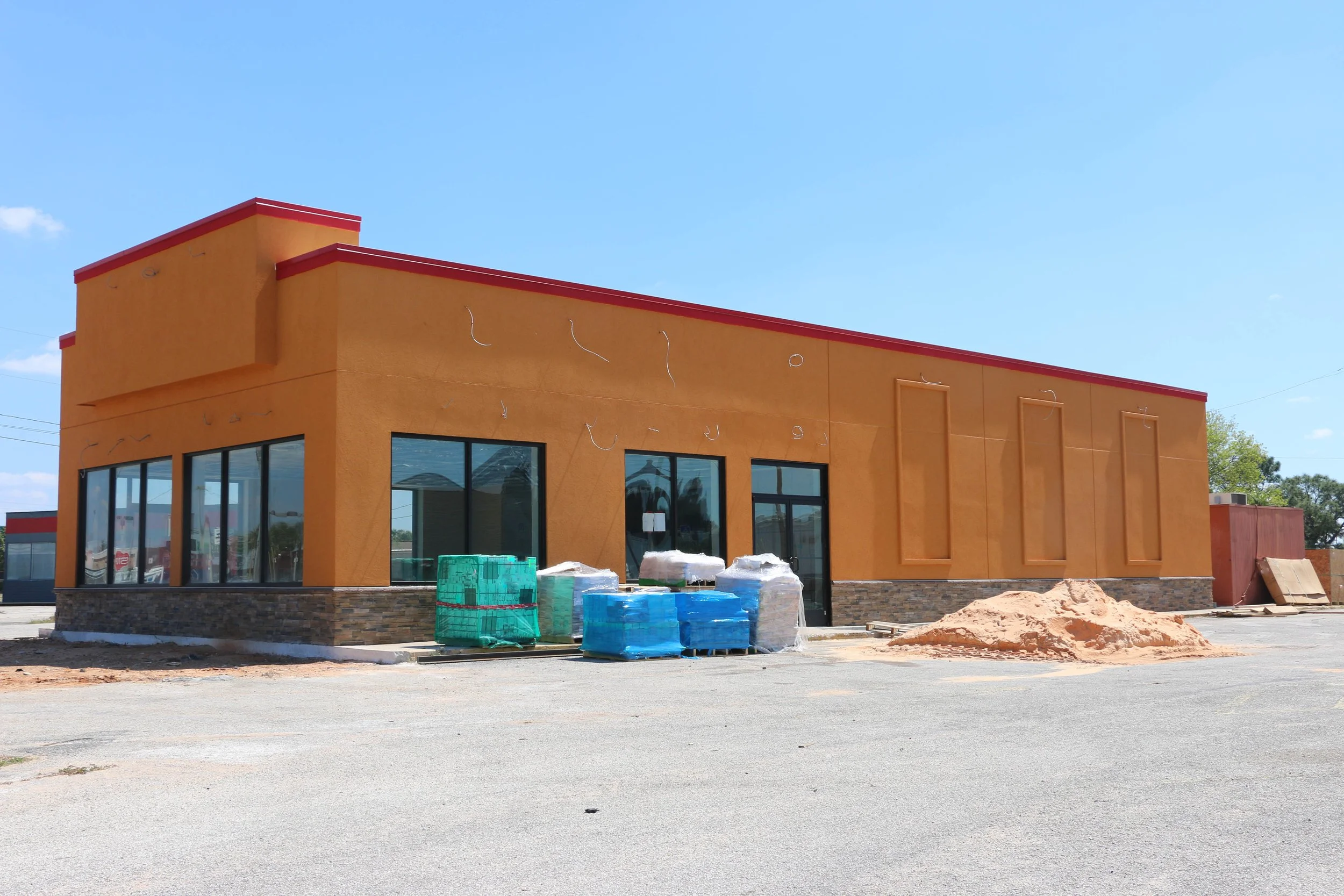 Under construction fast-food restaurant with orange walls, large windows, and red trim, construction materials and dirt pile in parking lot under blue sky