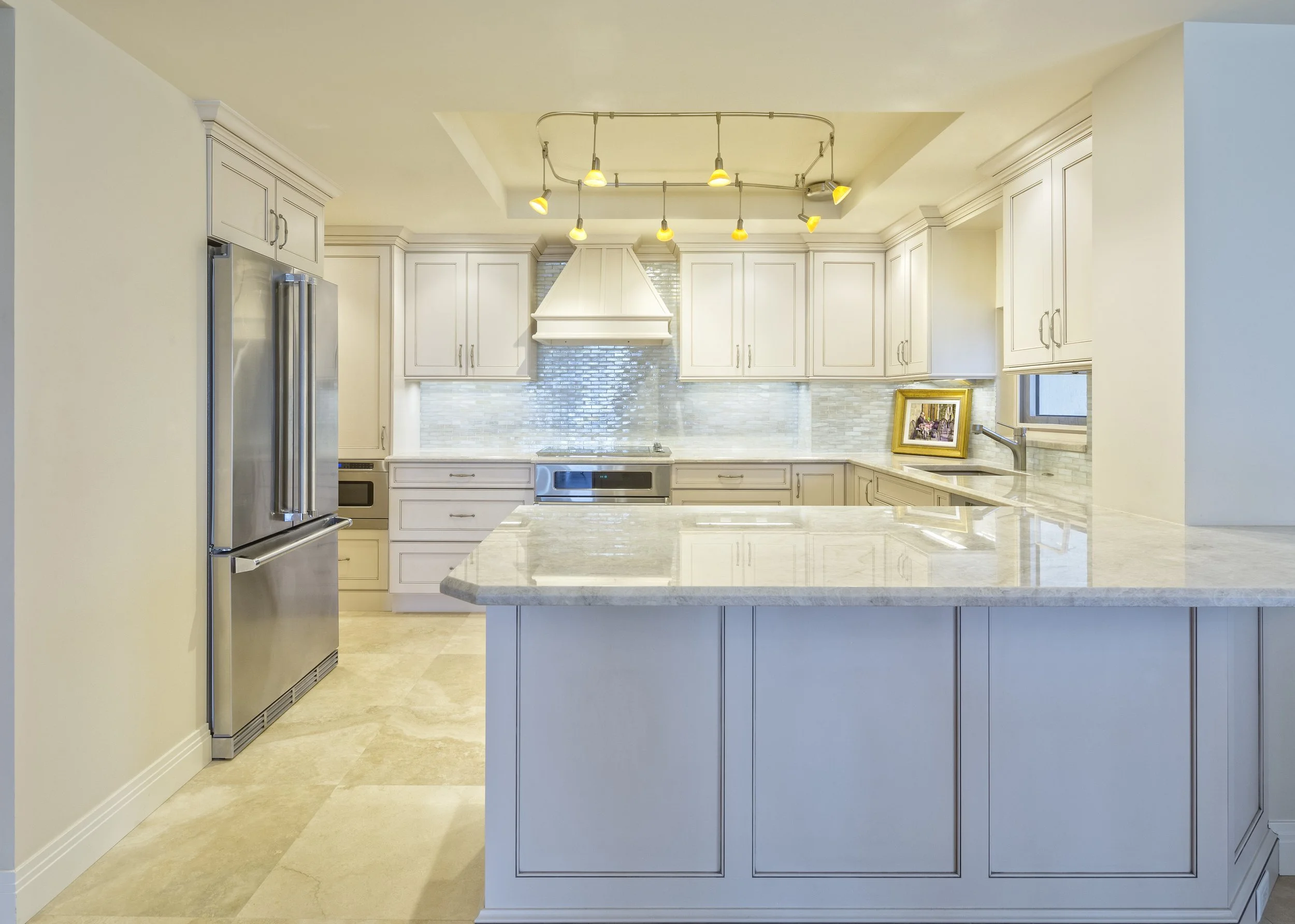 Modern white kitchen with marble countertops, stainless steel refrigerator, built-in oven, hood vent, and a window with a framed picture on the counter.