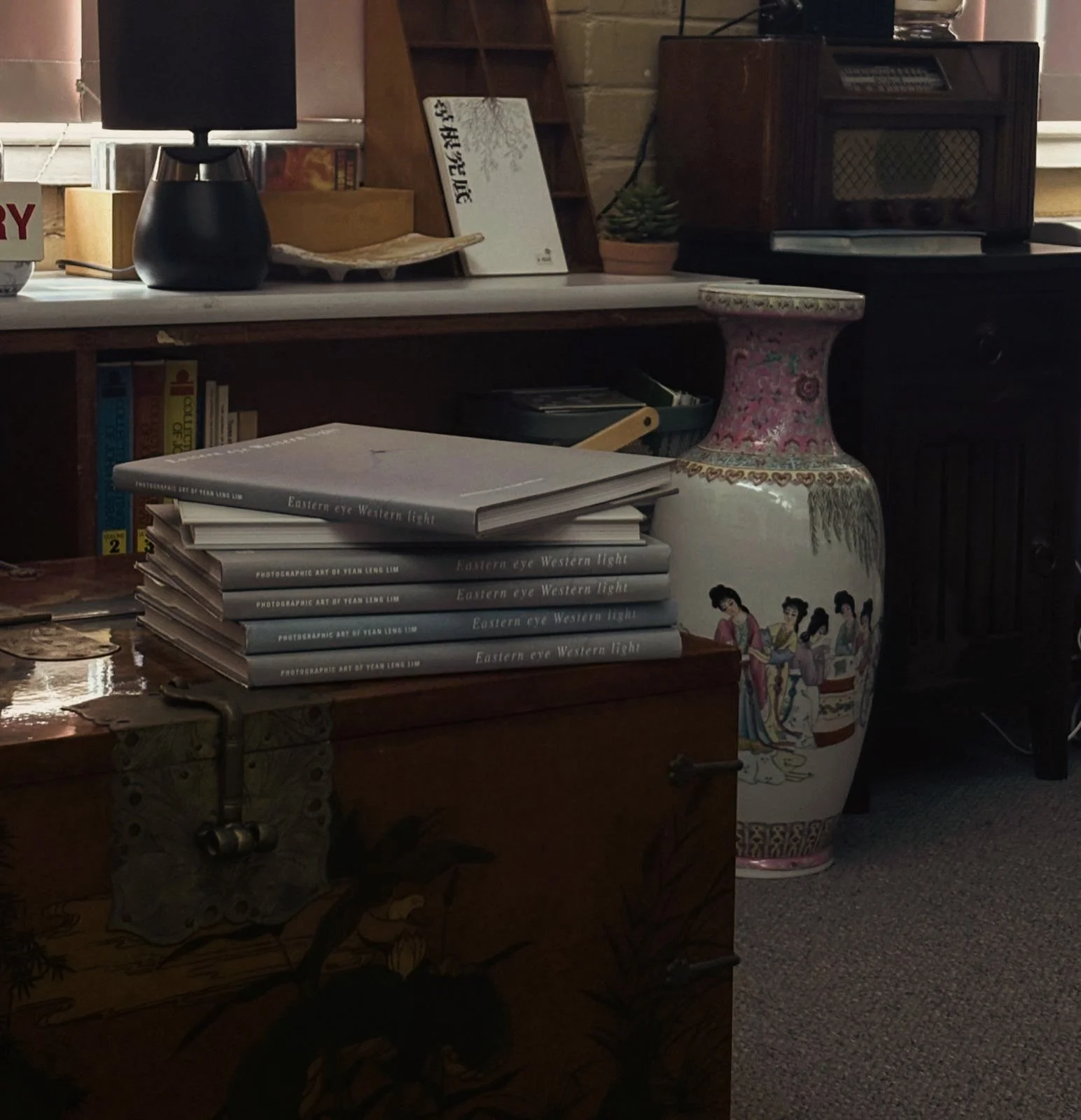 Stack of six books titled 'Eastern Eye Western Light' on a wooden surface, with a large decorative vase featuring traditional Asian artwork and by young women in colorful traditional dresses, a small lamp on a desk, a small plant, and various objects in the background.