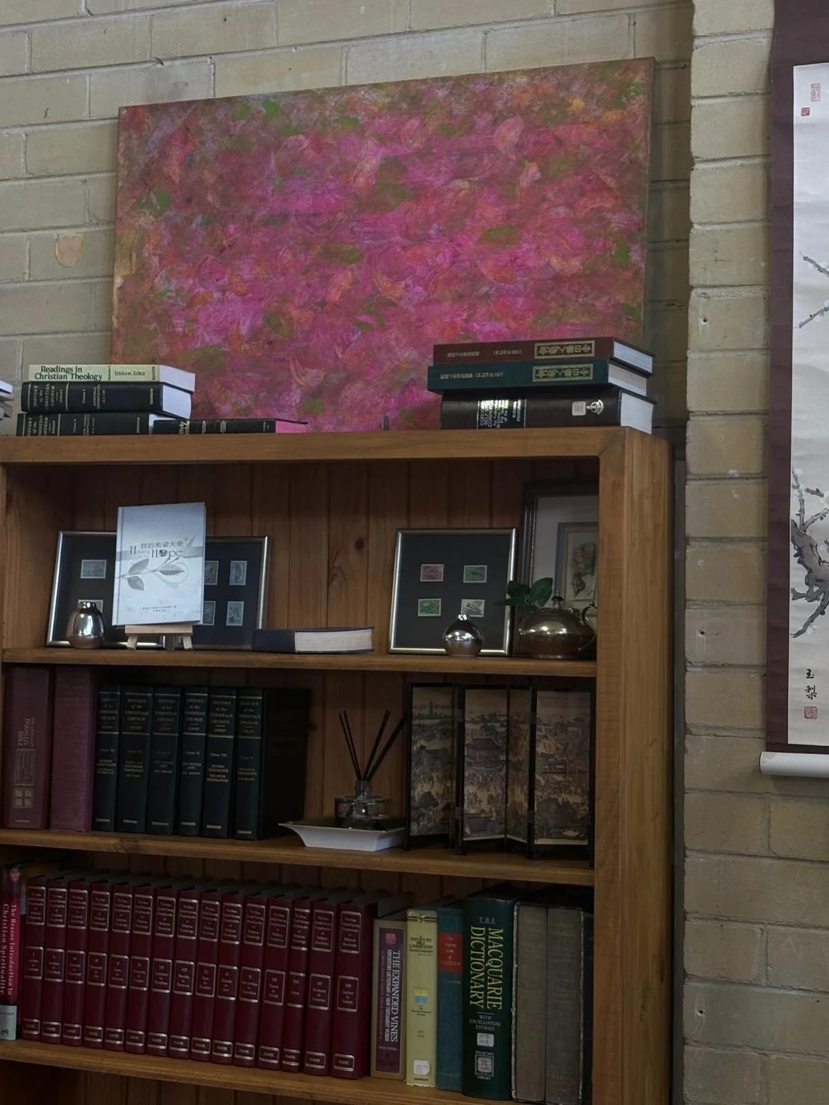 A wooden bookshelf against a brick wall with framed photos, books, and decorative objects. Behind the bookshelf, there is a large abstract painting with pink, purple, and green colors.