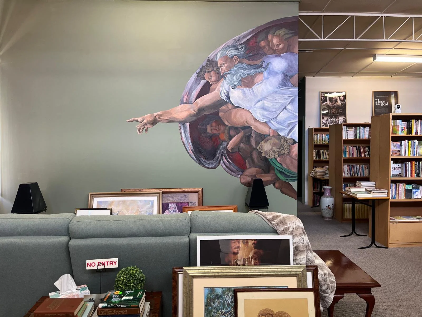Interior of a bookstore or library with a mural of God from the Sistine Chapel on the wall, bookshelves on the right, and a table with framed pictures and a