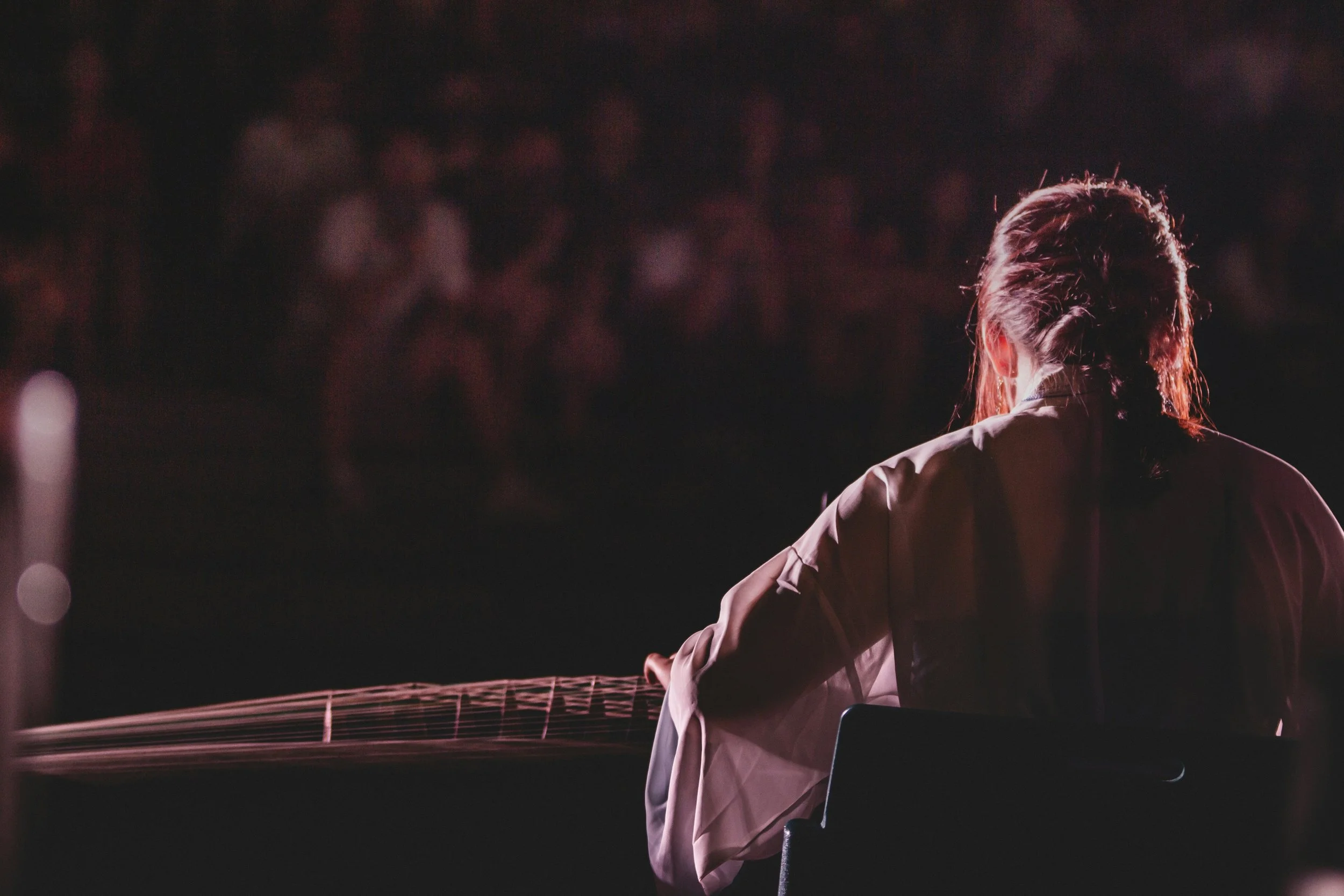 A woman with braided hair sitting outdoors at night, viewed from behind, with a guitar on her lap, and a blurry audience in the background.