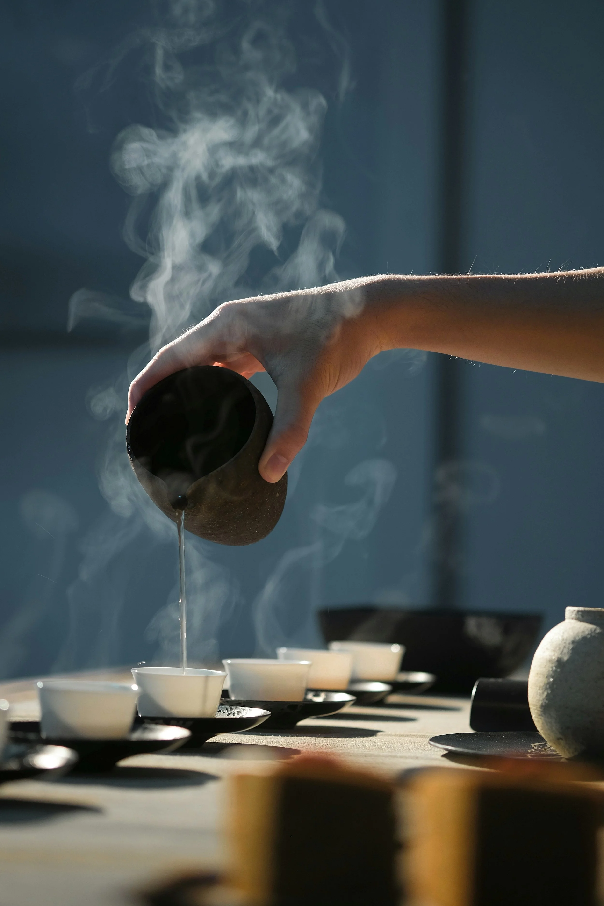 Person pouring hot tea into small white cups on black saucers, steam rising in a dimly lit room.