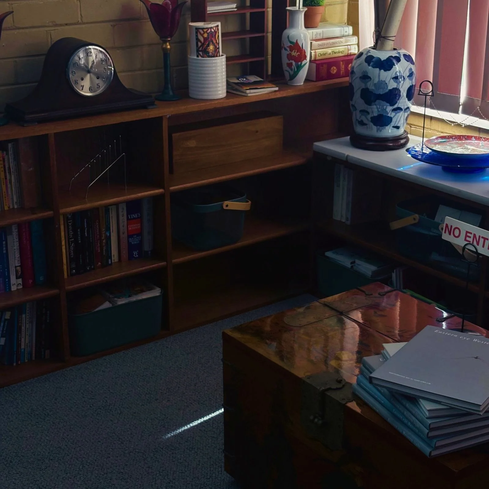 A cozy room with a wooden bookshelf filled with books, a vintage clock, decorative vases, and a table with stacked books. There's a window with pink curtains, and sunlight is partially illuminating the scene.