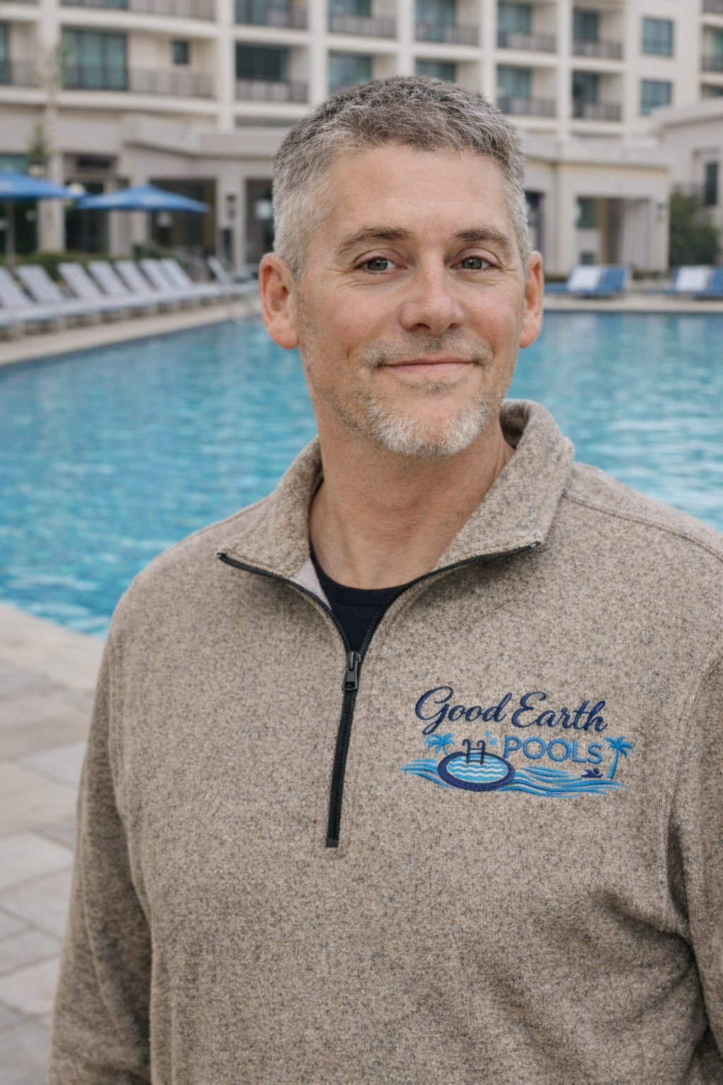 A man with short gray hair and a beard standing outdoors near a swimming pool in a residential complex, wearing a beige fleece jacket with the logo 'Good Earth Pools'.