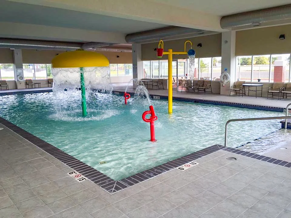 Commercial swimming pool with kid water features such as a mushroom shaped fountain, colorful water jets, and a yellow overhead water shower. There are lounge chairs around the pool and large windows in the background.Good Earth Pools, Oklahoma