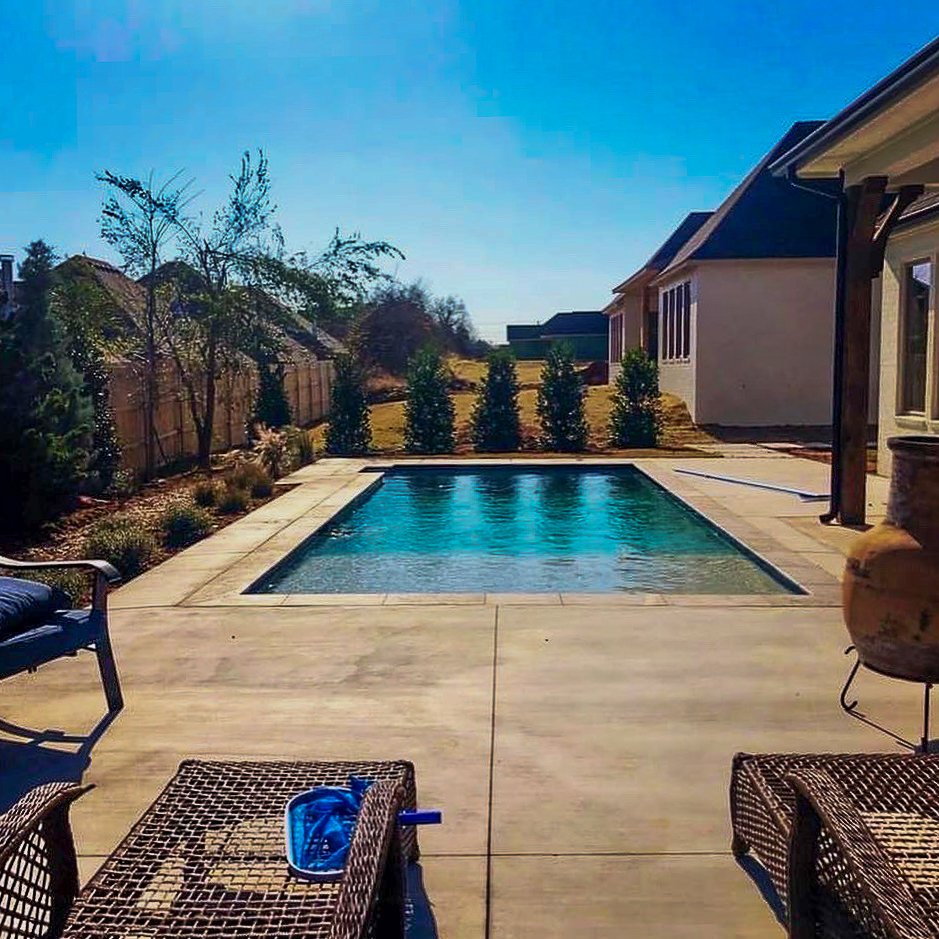 Custom rectangular swimming pool, surrounded by a concrete deck, with some patio furniture, a hedge of small trees, and a house in the background under a clear blue sky. Good Earth Pools, Oklahoma