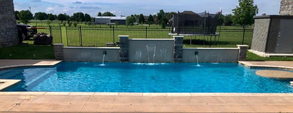 In-ground backyard swimming pool with water features, surrounded by a concrete deck, with a fence, grassy field, and open sky in the background.