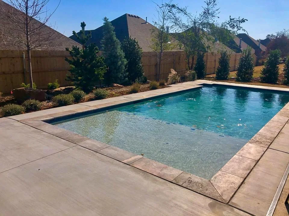 Backyard swimming pool with a concrete deck and a landscaped fence line with bushes and trees. Good Earth Pools, Oklahoma