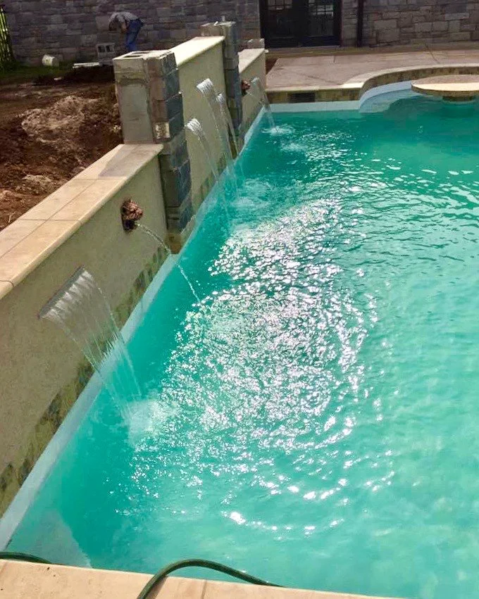 Swimming pool with three small waterfalls flowing into it, surrounded by tan tiles and a stone wall in the background.