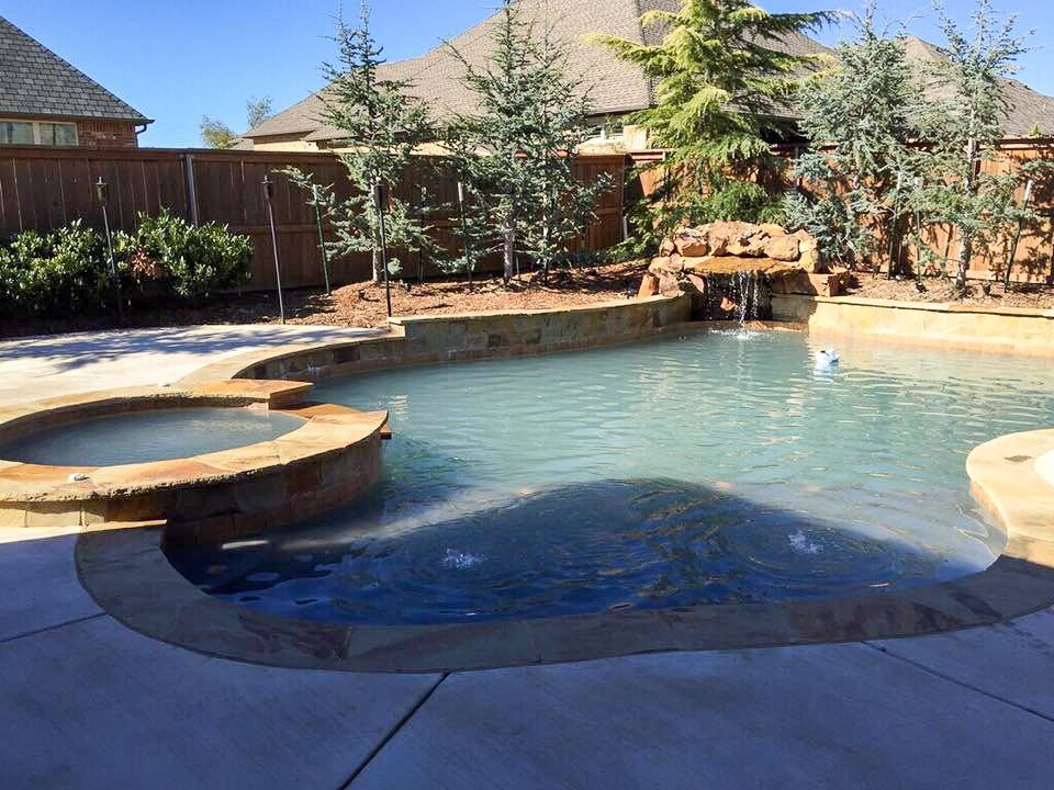 Custom swimming pool, hot tub, and waterfall feature, surrounded by a concrete patio, trees, and a wooden fence. Good Earth Pools, Oklahoma