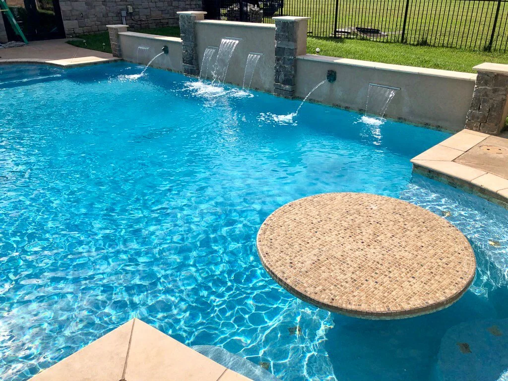 Custom swimming pool table top, bar, water features with streams flowing into the pool, and a grassy yard with a fence in the background. Good Earth Pools, Oklahoma. 