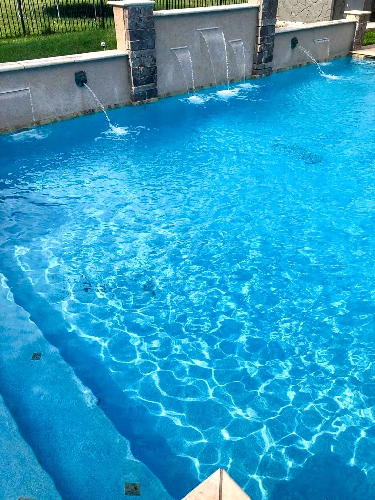 Custom residential swimming pool with waterfall features built into a stone and concrete wall, with water flowing from three spouts into the pool. There is grass and a black fence in the background.