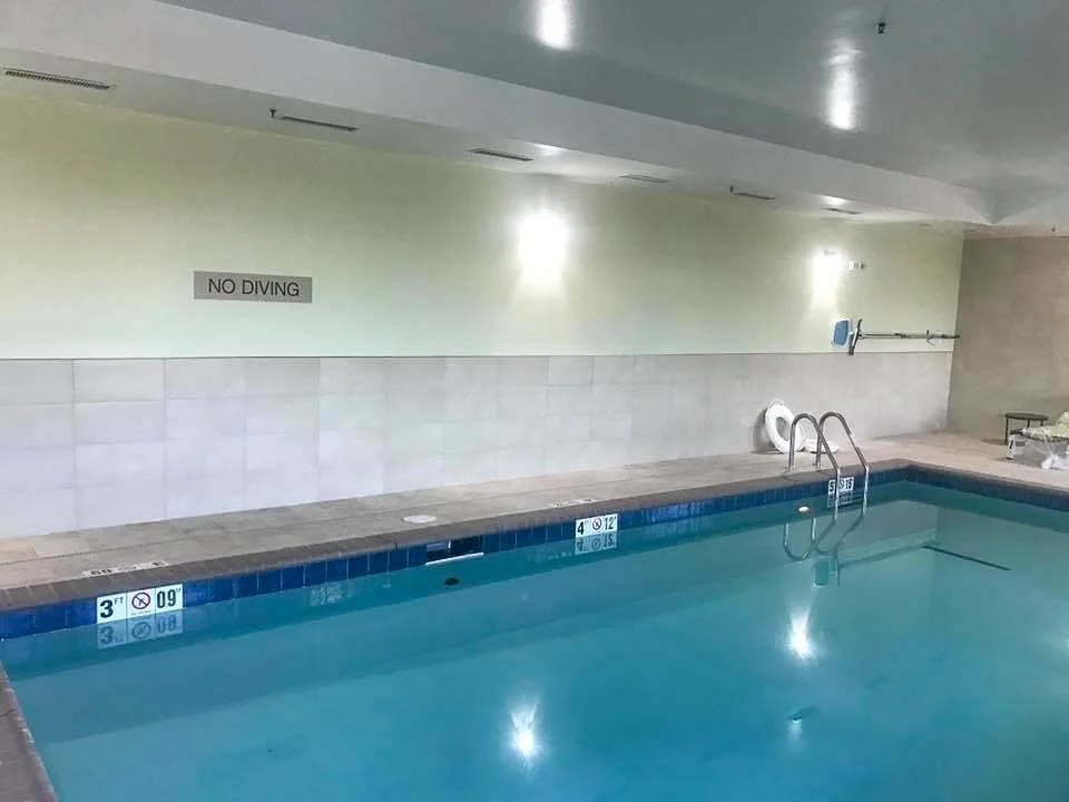 Commercial indoor swimming pool with a no diving sign on the wall, pool depth markers 3 feet and 4 feet, metal handrails at the pool steps, and pool cleaning equipment on the wall.