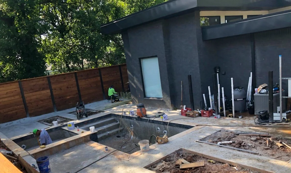 Construction workers building a pool in the backyard of a modern house with a dark exterior, surrounded by trees and a wooden fence. Good Earth Pools, Oklahoma