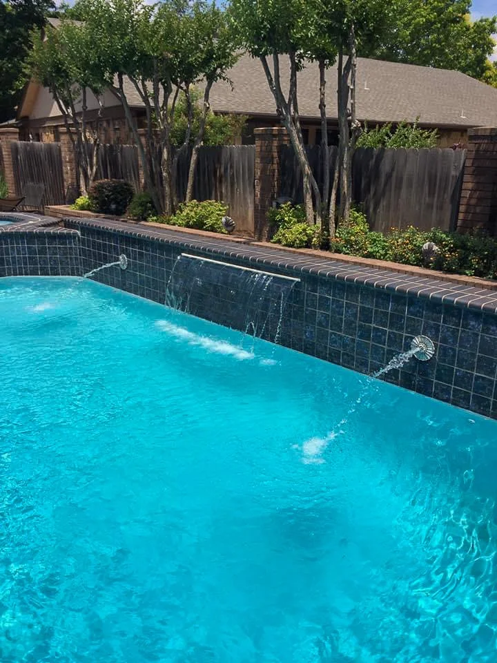 Custom swimming pool with waterfalls, surrounded by trees, bushes, and a wooden fence, in a backyard. Good Earth Pools, Oklahoma