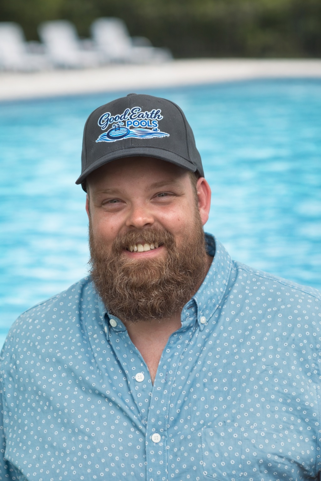 A smiling man with a beard wearing a blue patterned shirt and a cap that says 'Good Earth Pools', standing near a swimming pool with lounge chairs in the background.
