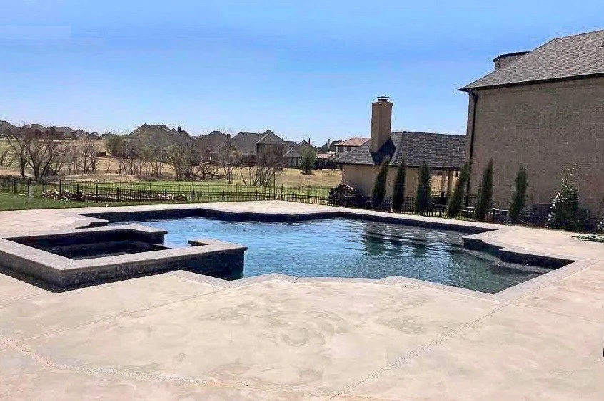 Custom swimming pool with a hot tub, surrounded by a concrete patio. There are houses and trees in the background under a blue sky. Good Earth Pools, Oklahoma