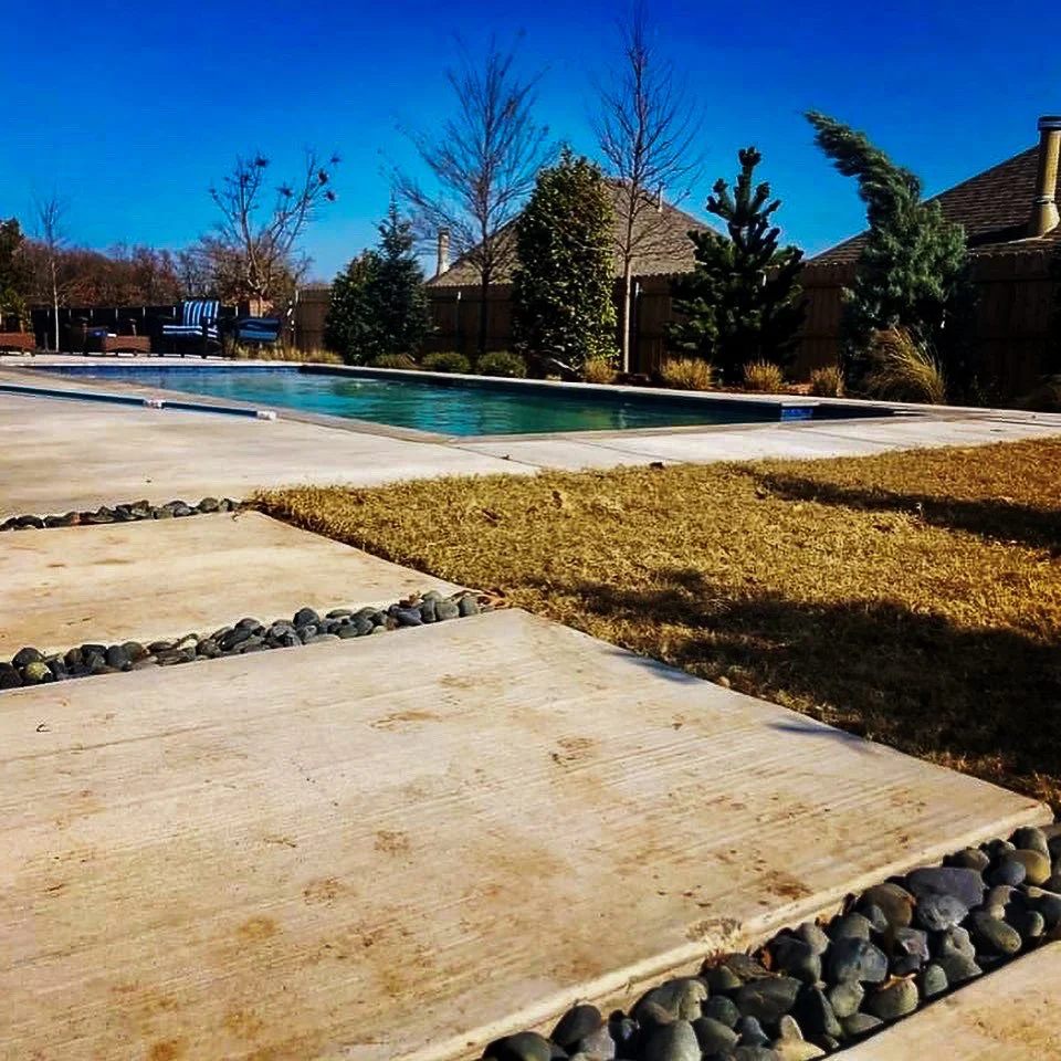 Custom swimming pool, paved patio, grass, trees, wooden fence, and a house roof visible under a clear blue sky. Good Earth Pools, Oklahoma