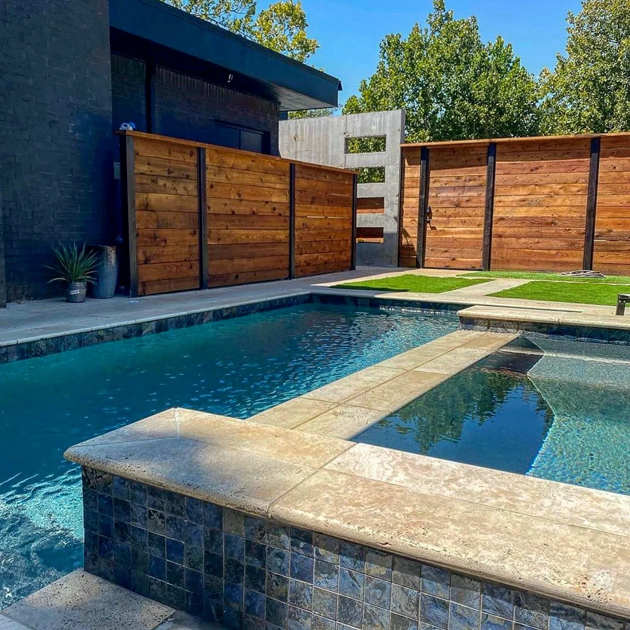 Custom swimming pool, a concrete pool deck, modern wooden privacy fences, and a lawn area with artificial grass. There are trees and a house with a dark exterior in the background. Good Earth Pools, Oklahoma